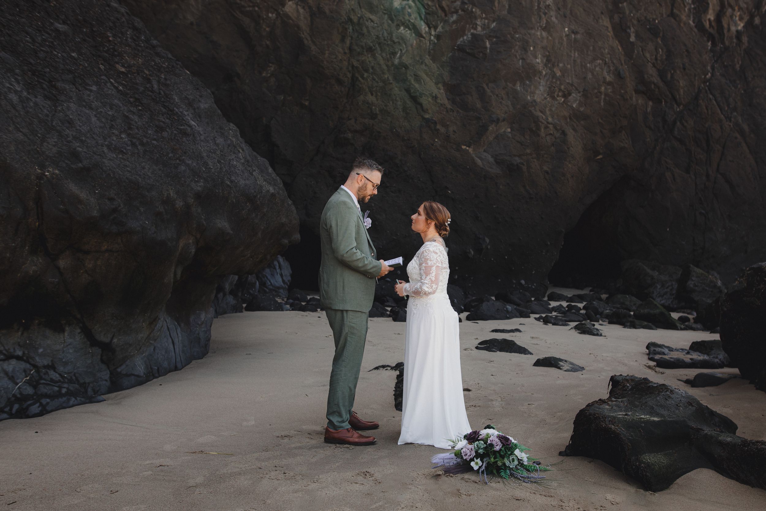 A couple in wedding attire shares an intimate moment on a dramatic black sand beach surrounded by rocky cliffs.