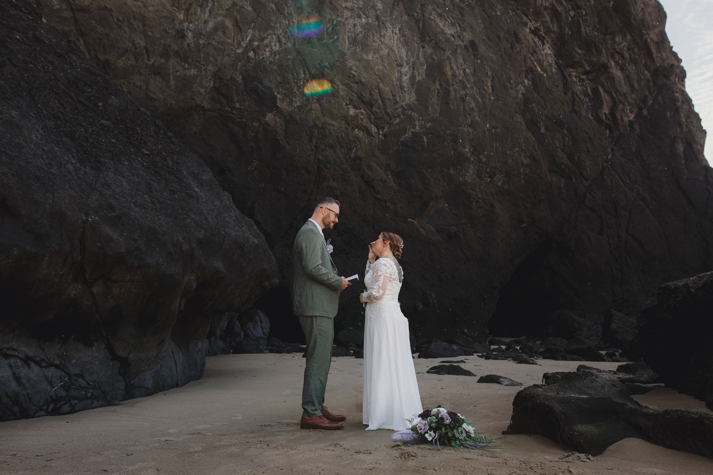 Romantic coastal wedding ceremony with couple standing between dramatic rock formations on sandy beach.