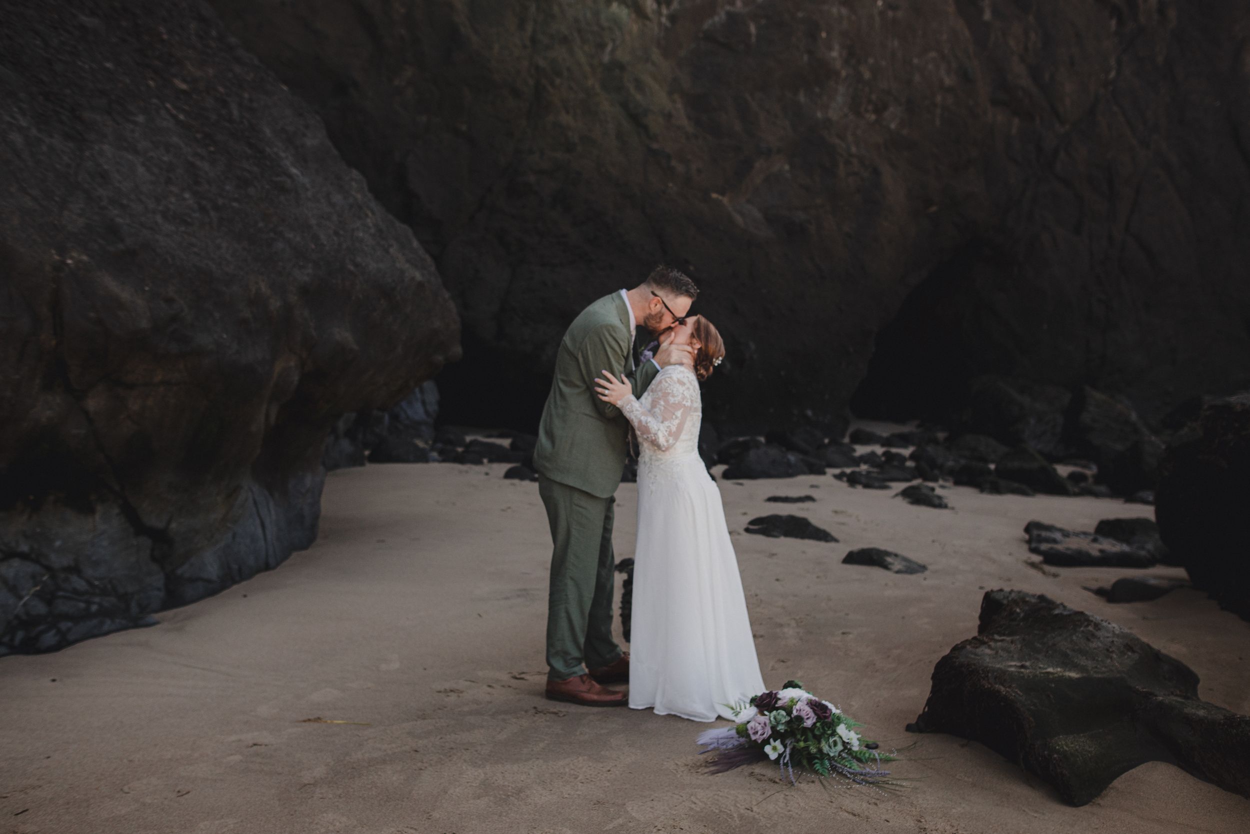 Intimate beach wedding scene with couple sharing tender moment among dark coastal rock formations.