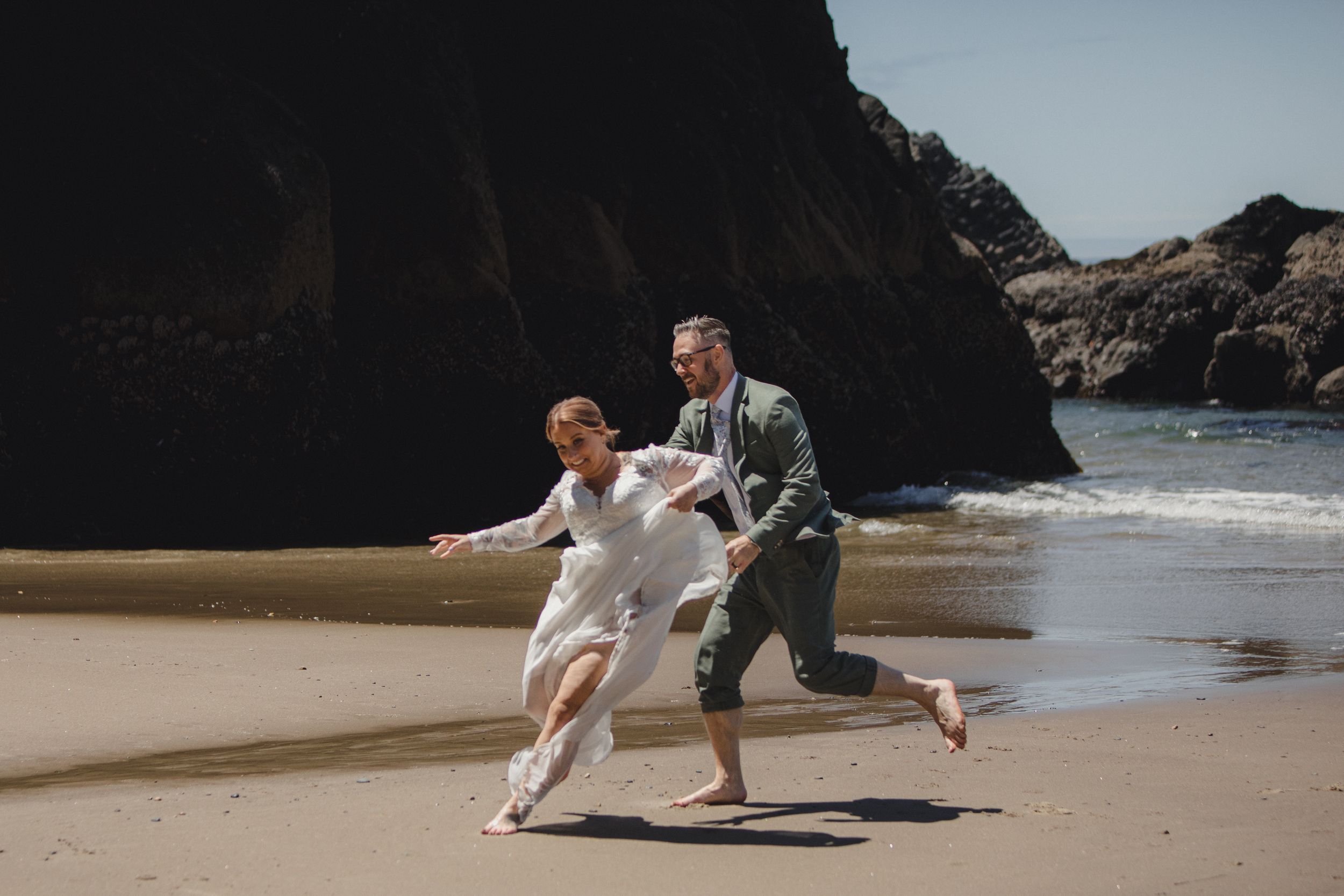 A couple runs playfully along a scenic beach with rocky cliffs in the background on their wedding day.