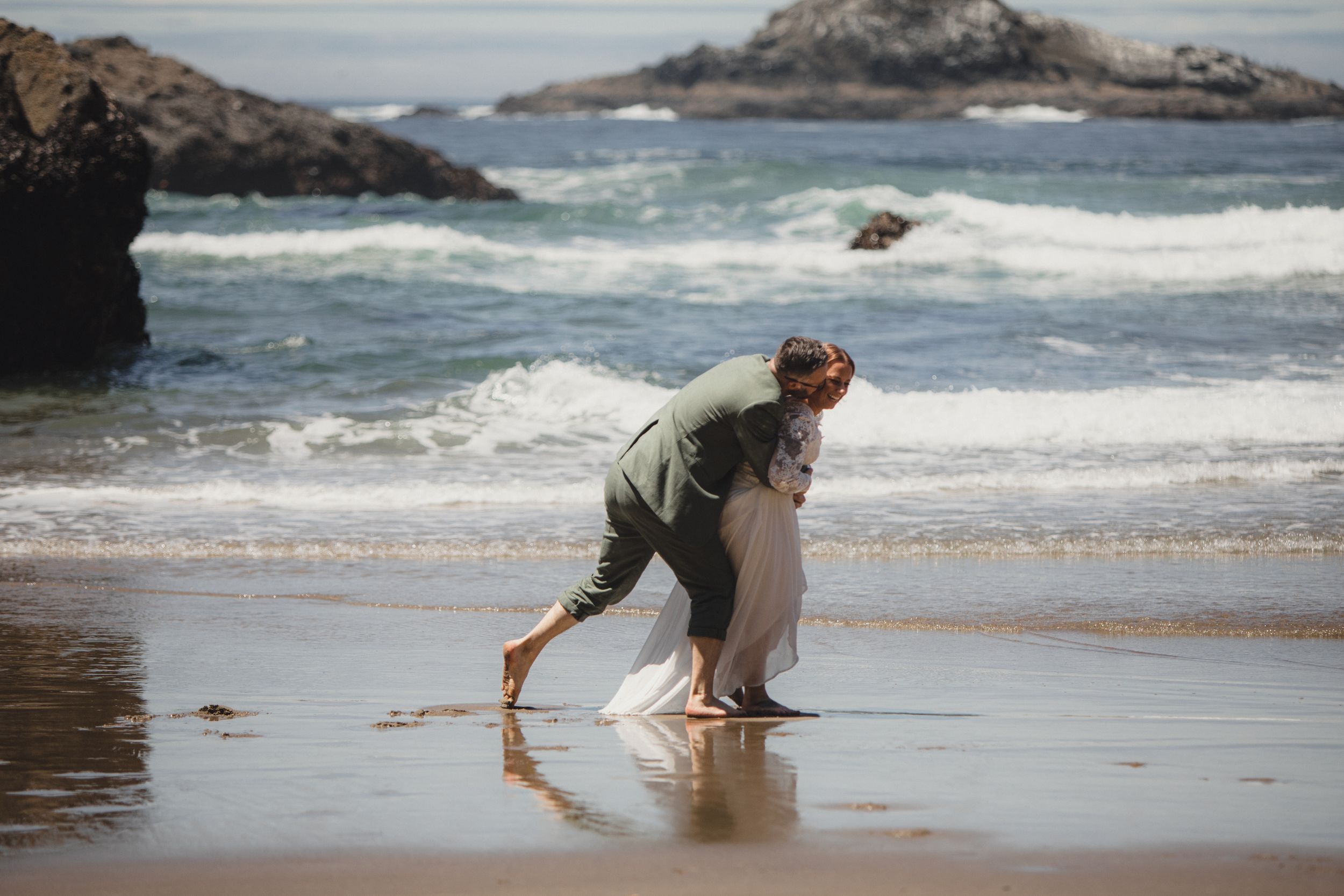 Person in waders searches for clams at low tide along a scenic rocky beach with crashing waves.