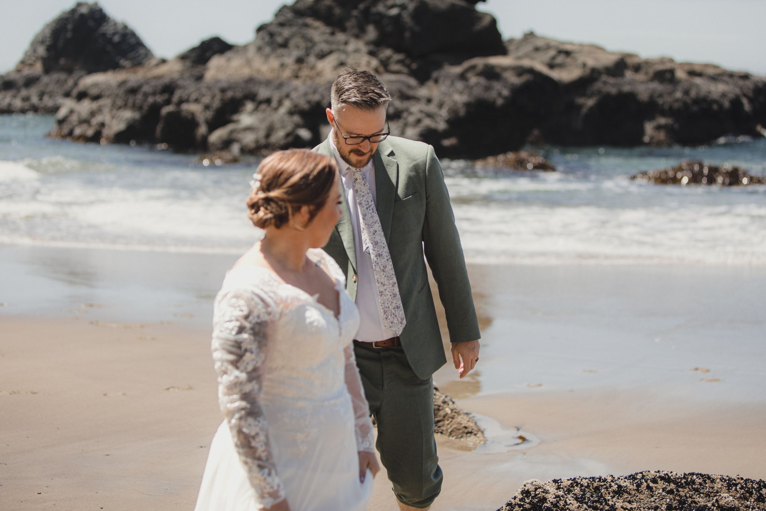 A couple shares romantic moments along a rocky beach coastline in elegant wedding attire.