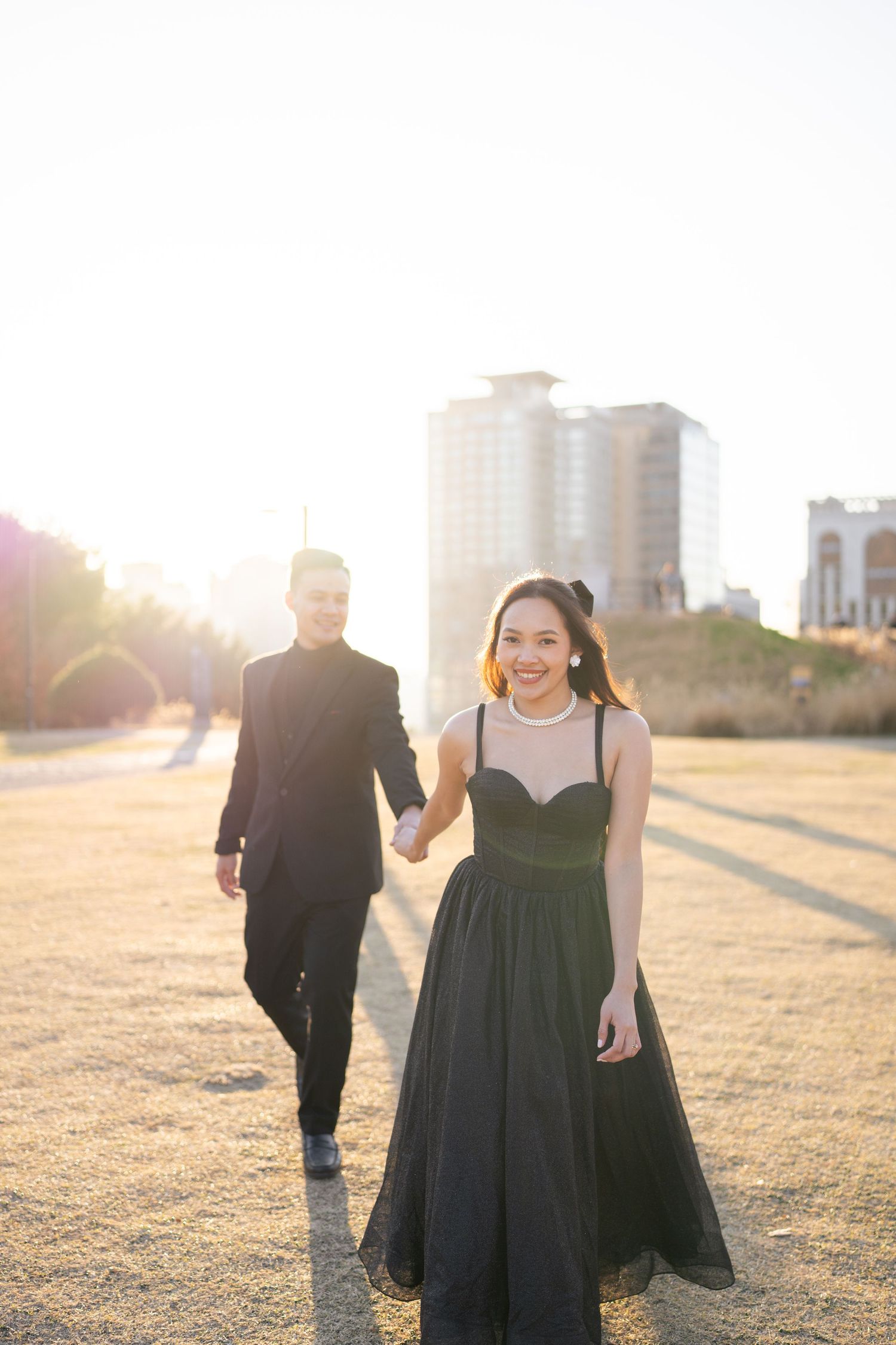 A couple holds hands while walking in a sunlit field at golden hour, with the woman wearing a flowing dark dress.