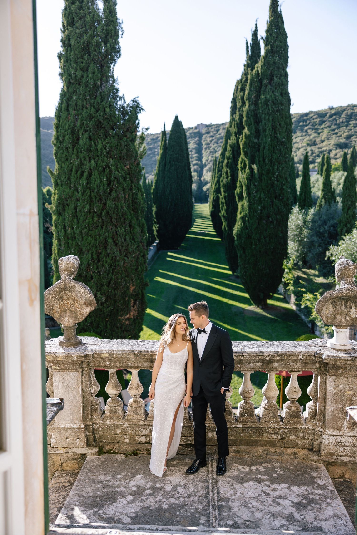 Elegant wedding couple stands on stone terrace with cypress trees and scenic Tuscan landscape in background.