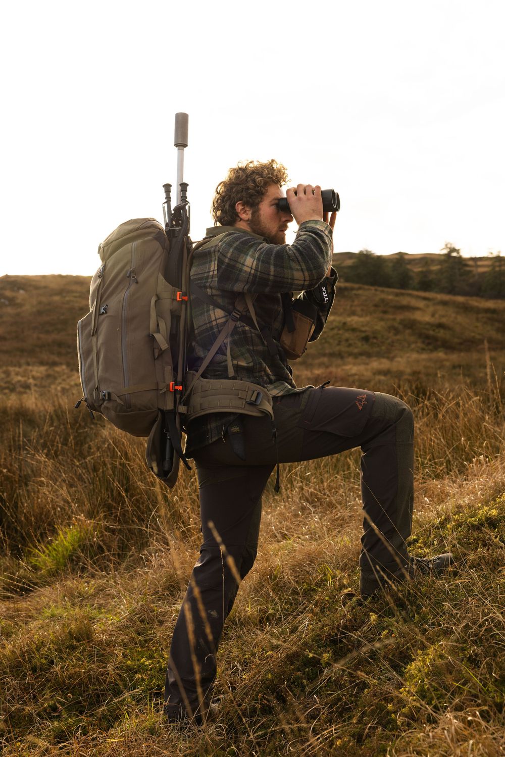 Person with backpack and binoculars observes wildlife while standing in golden grassland at sunset.