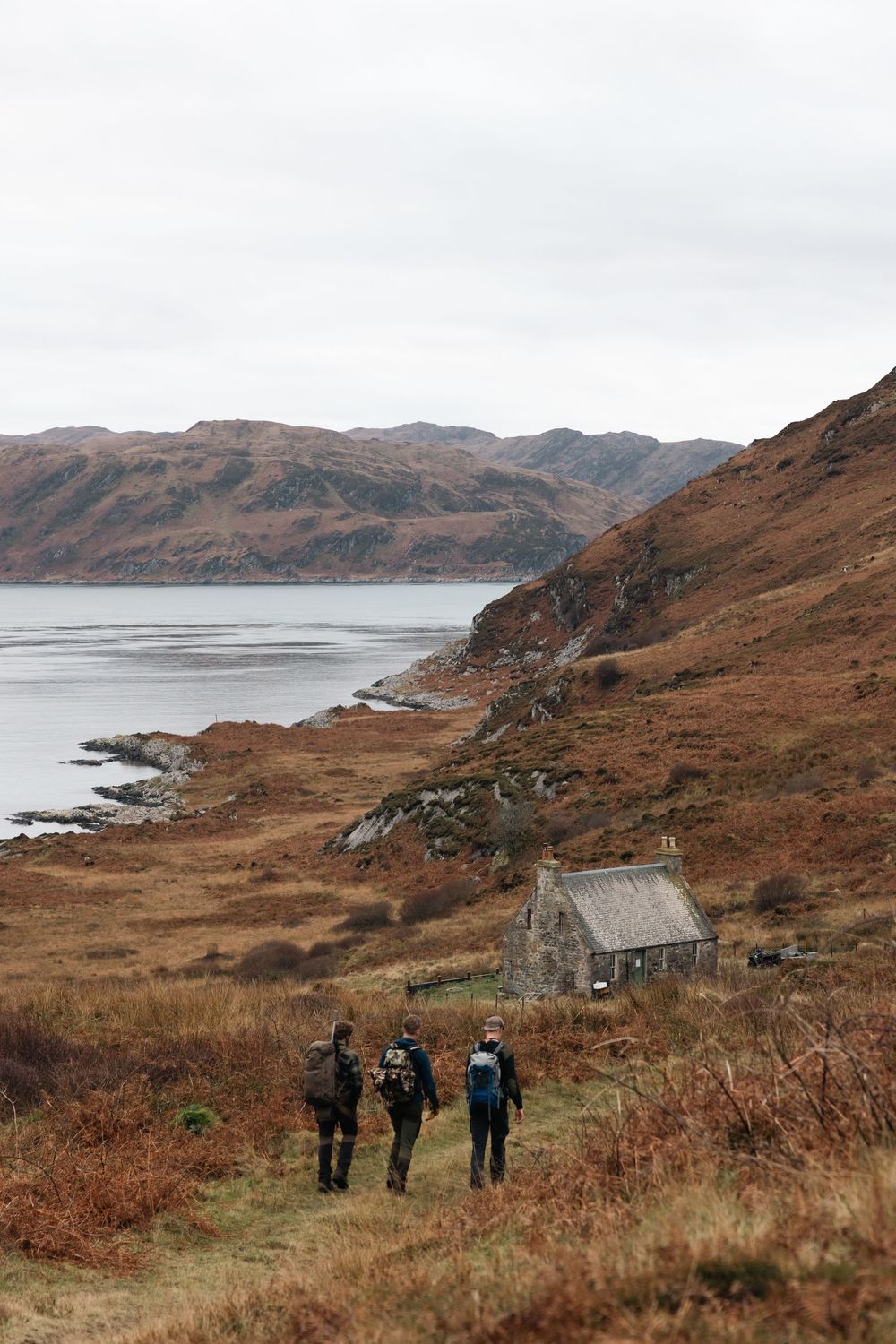 Hikers trek along a rugged coastal trail in Scotland with mountains and a stone cottage in the scenic landscape.