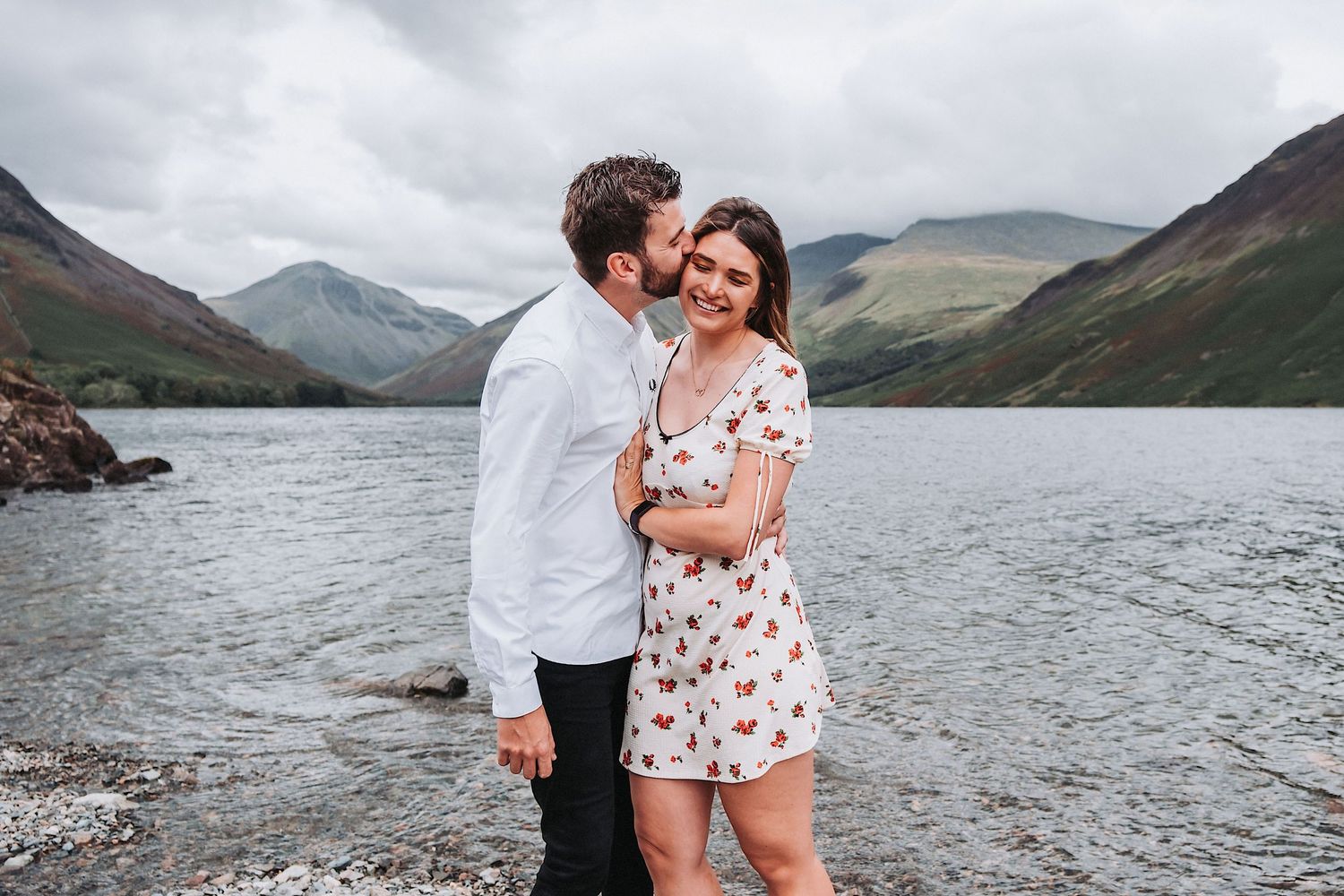 A couple embraces by a scenic mountain lake with rocky shores and misty peaks in the background.
