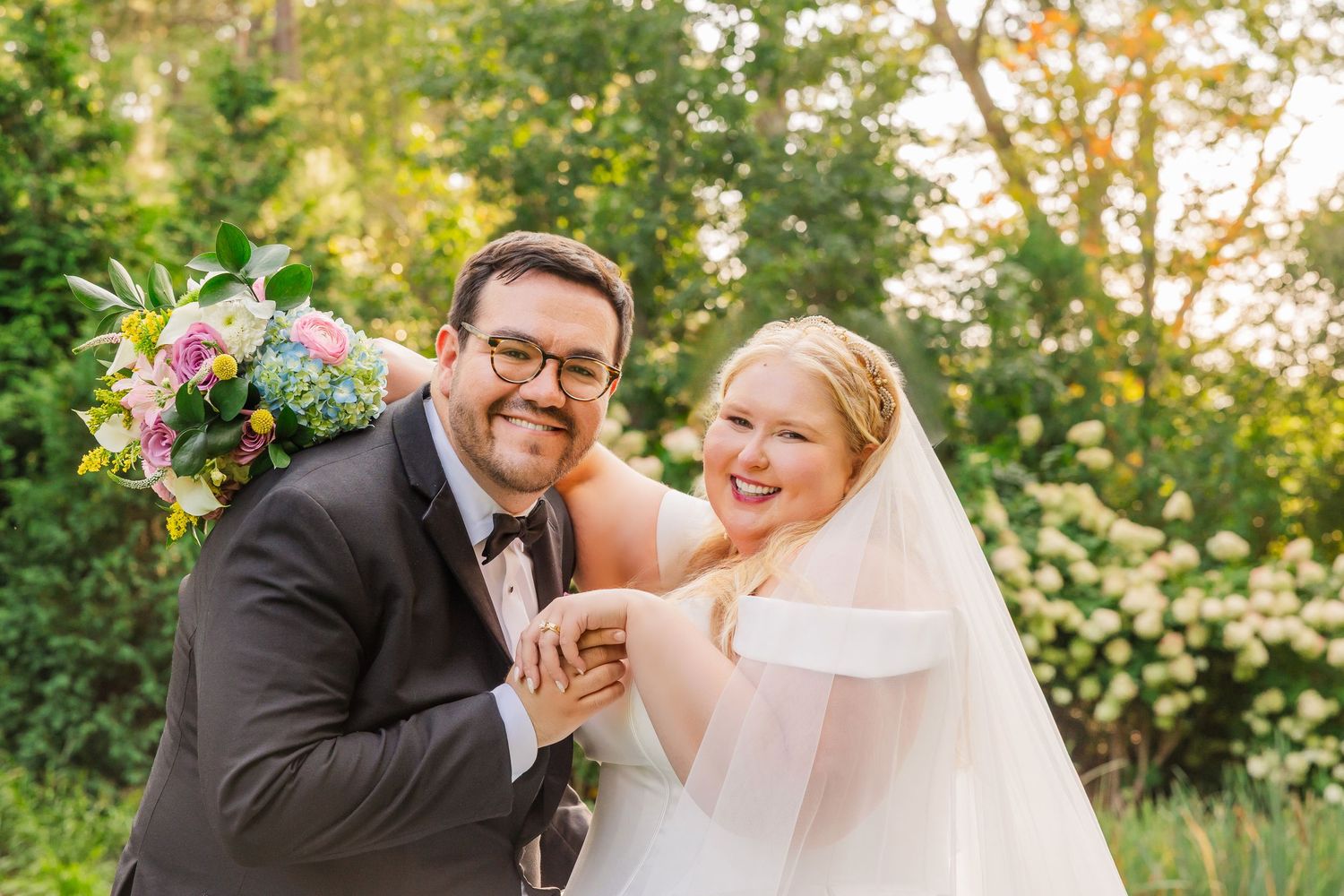 A joyful wedding couple laughs together outdoors while holding a colorful floral bouquet in golden sunlight.
