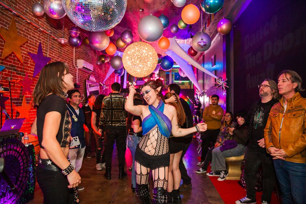 People dancing and socializing at a nightclub with colorful purple lighting and disco balls overhead.