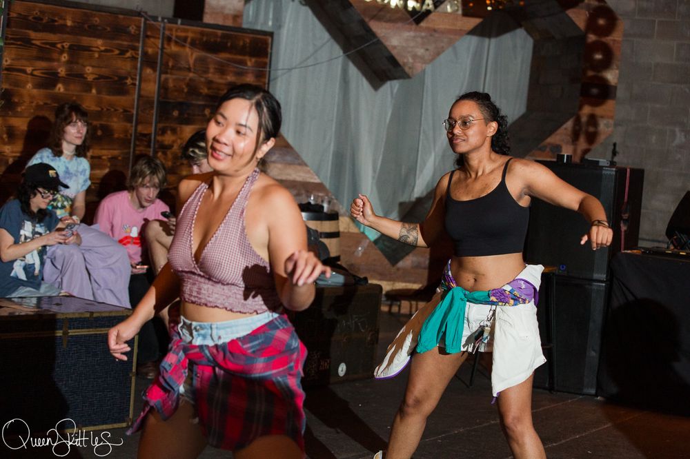 Two women dancing energetically at an indoor event, wearing colorful workout attire with tied scarves around their waists.