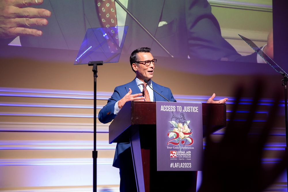 A speaker gestures while presenting at a business conference podium with purple stage lighting in the background.