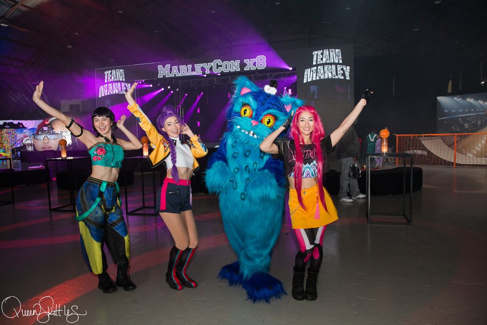 Four costumed performers pose with a blue monster mascot at MarleyCon xB event on a colorful lit stage.