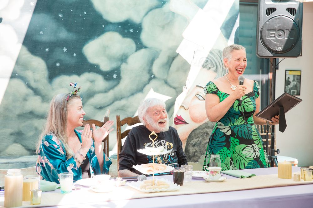 Three people celebrate at a party with cake and drinks while laughing together at a decorated table.