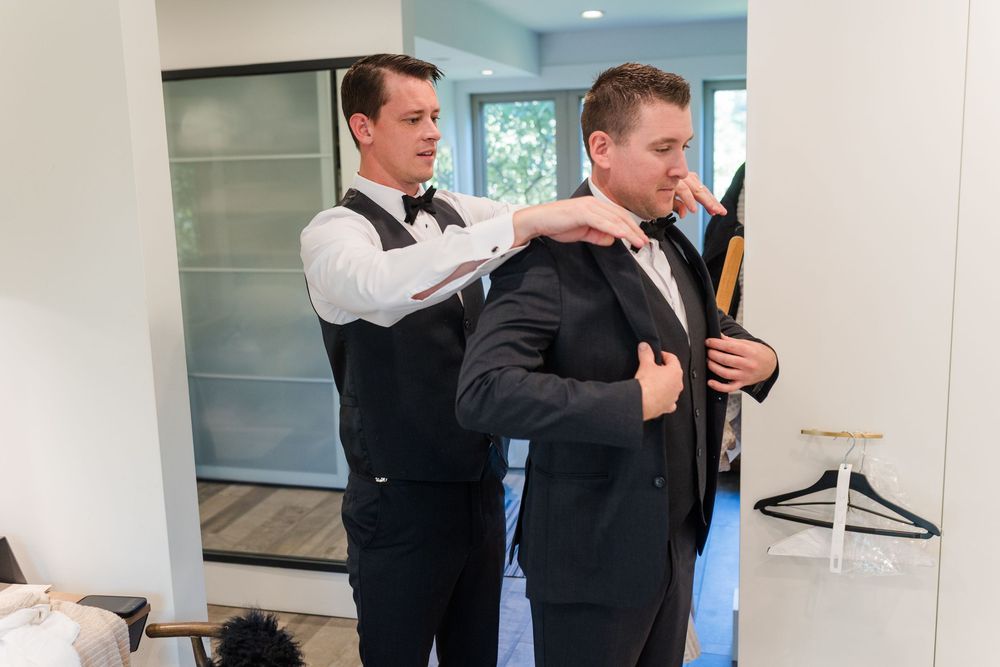 Two people in formal attire adjust their bowties while standing in a bright interior space.