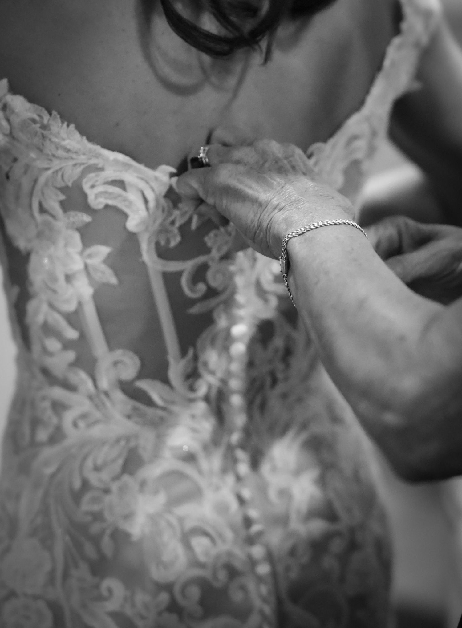 Black and white photo of a bride in lace wedding gown being buttoned up by an older woman wearing a bracelet.