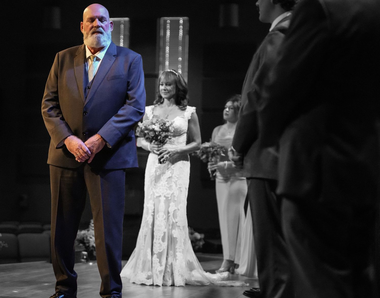 Bride in white lace gown walks down aisle with older man in blue suit at elegant wedding ceremony.