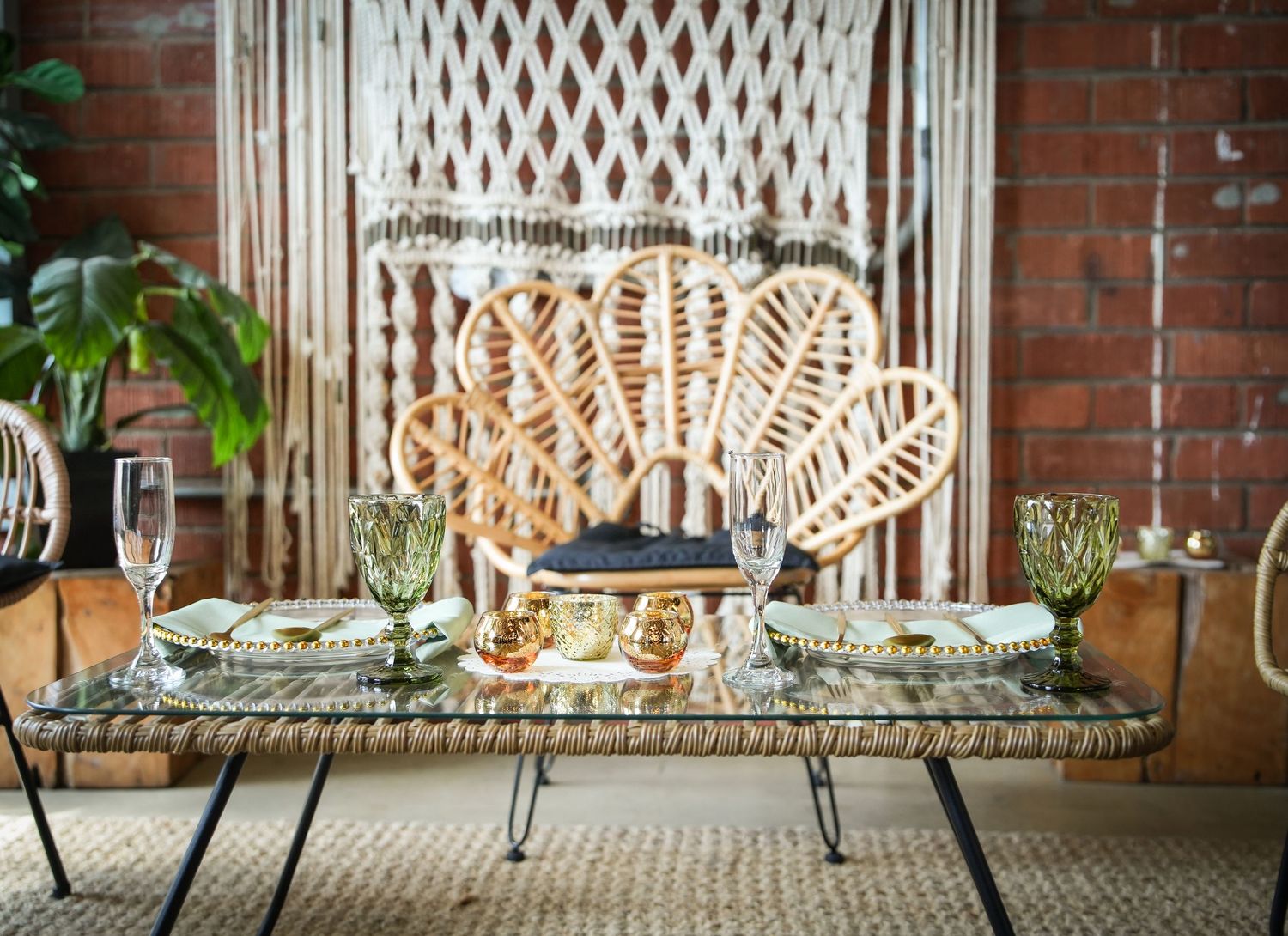 Boho wedding tablescape with rattan peacock chair, macrame backdrop, glass table, green goblets, and gold accents on jute rug.