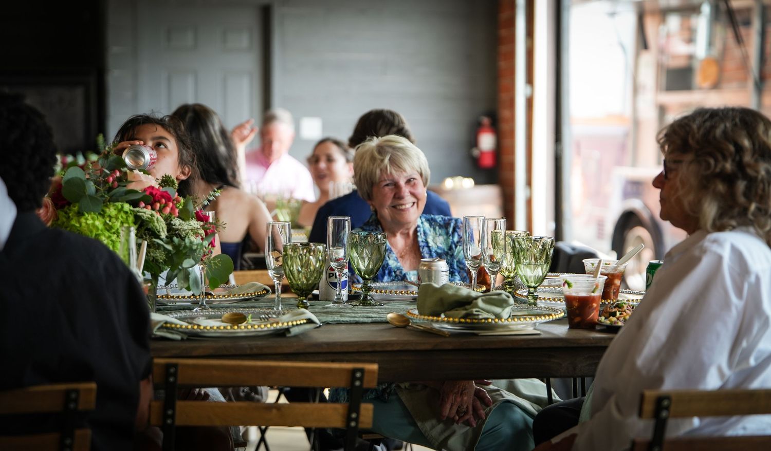 Guests smiling and laughing at a beautifully set dinner table with floral centerpieces in a rustic restaurant.