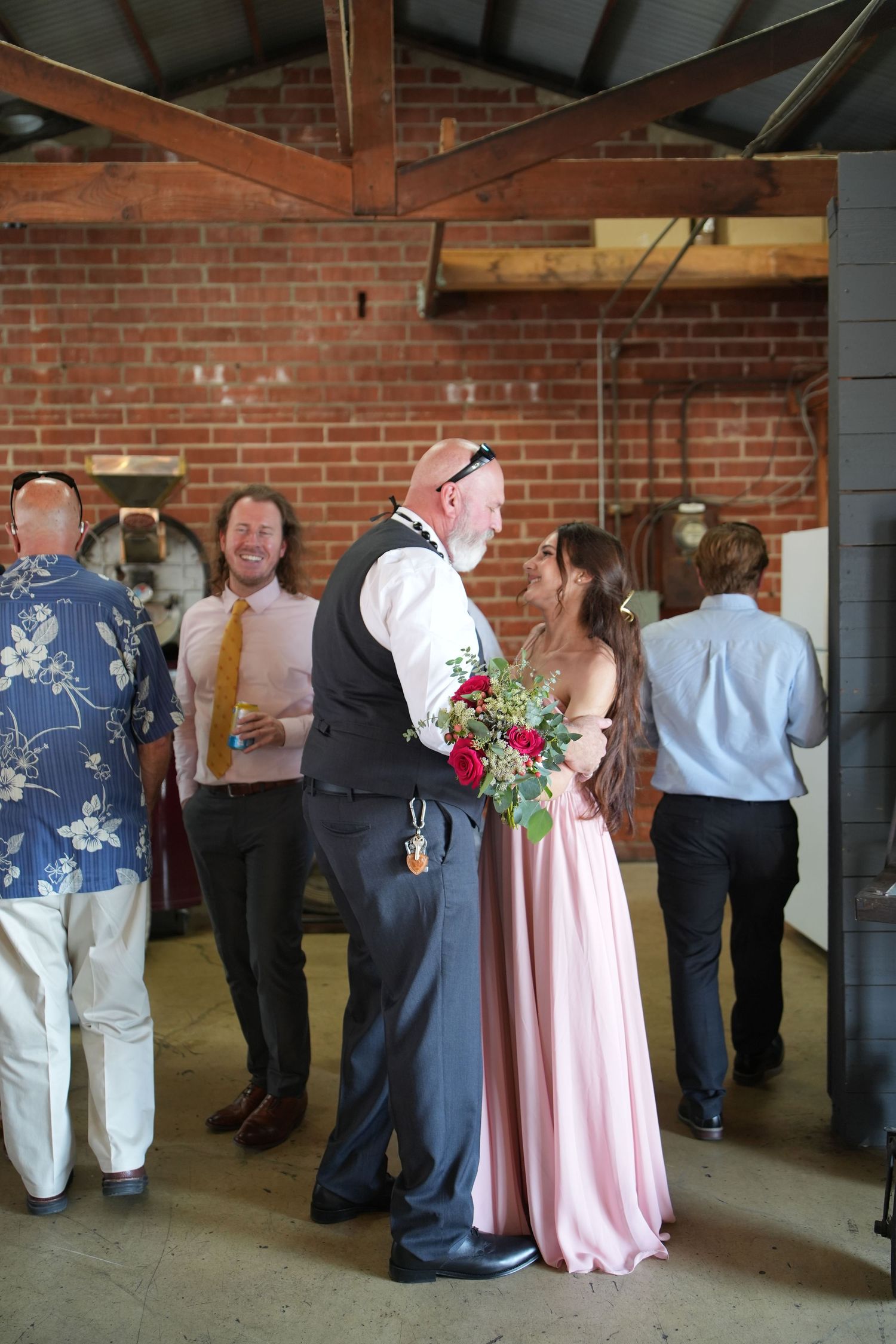 Bride in pink gown holds red bouquet while sharing a moment with groom in rustic brick venue.