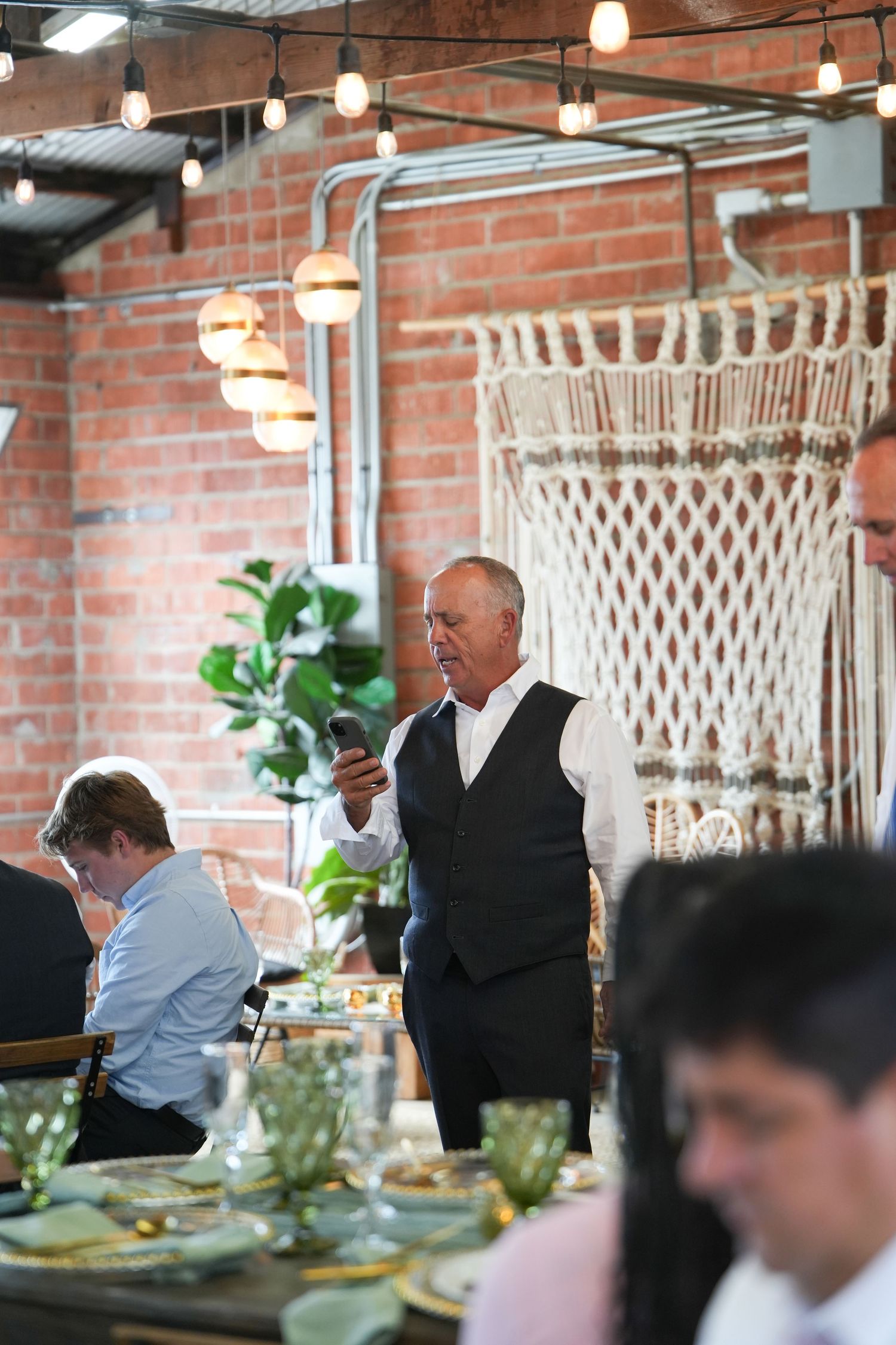 Man in vest checks phone at elegant indoor wedding reception with brick walls and macrame decor.