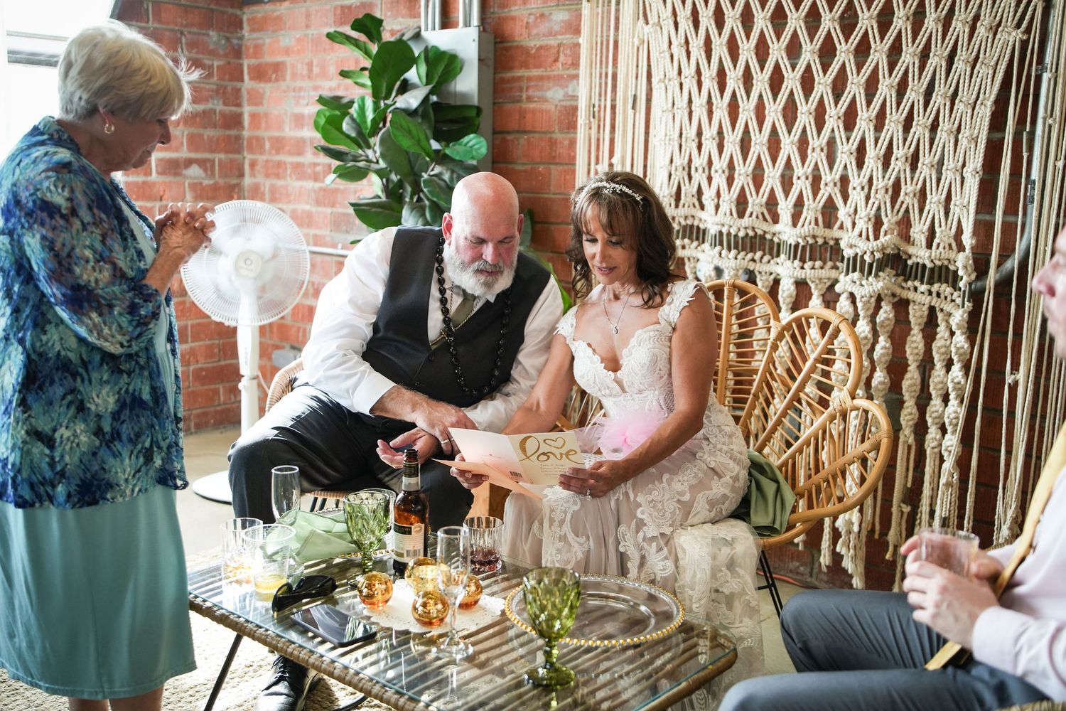 Bride and groom reviewing wedding vows seated together, officiant nearby, boho decor with macrame backdrop and brick walls.