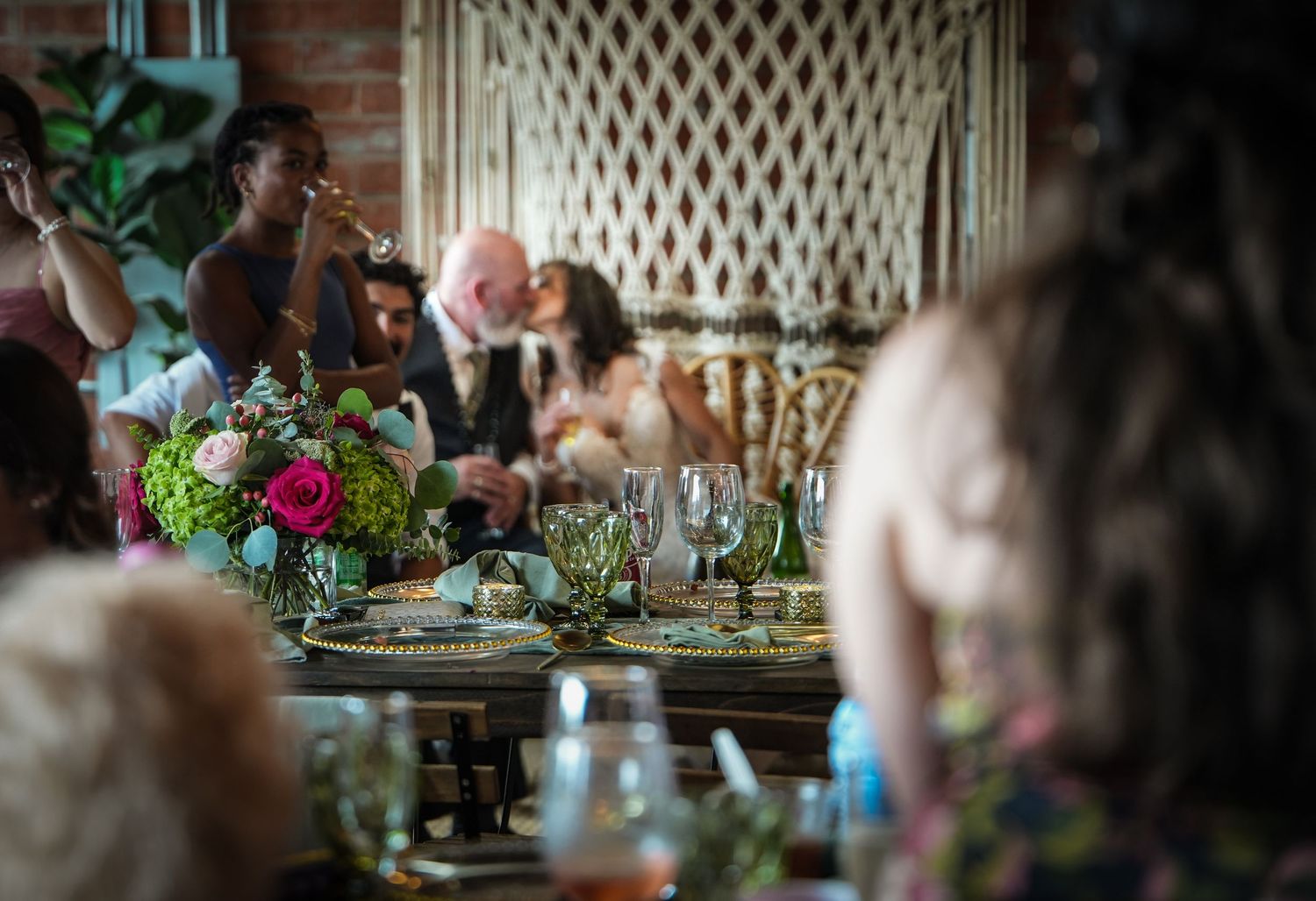 Elegant wedding reception dinner table with floral centerpieces, glassware, and guests socializing in a warmly lit venue.
