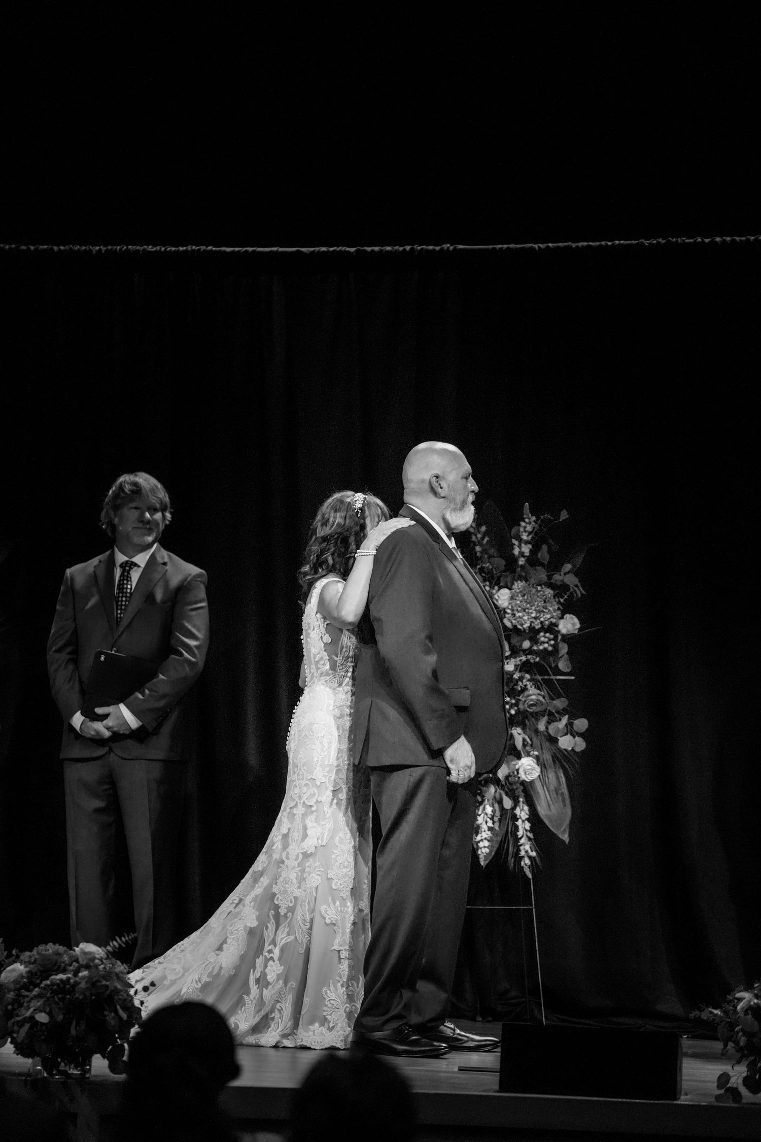 Black and white wedding ceremony photo of bride embracing older man, with officiant standing nearby and floral arrangement.