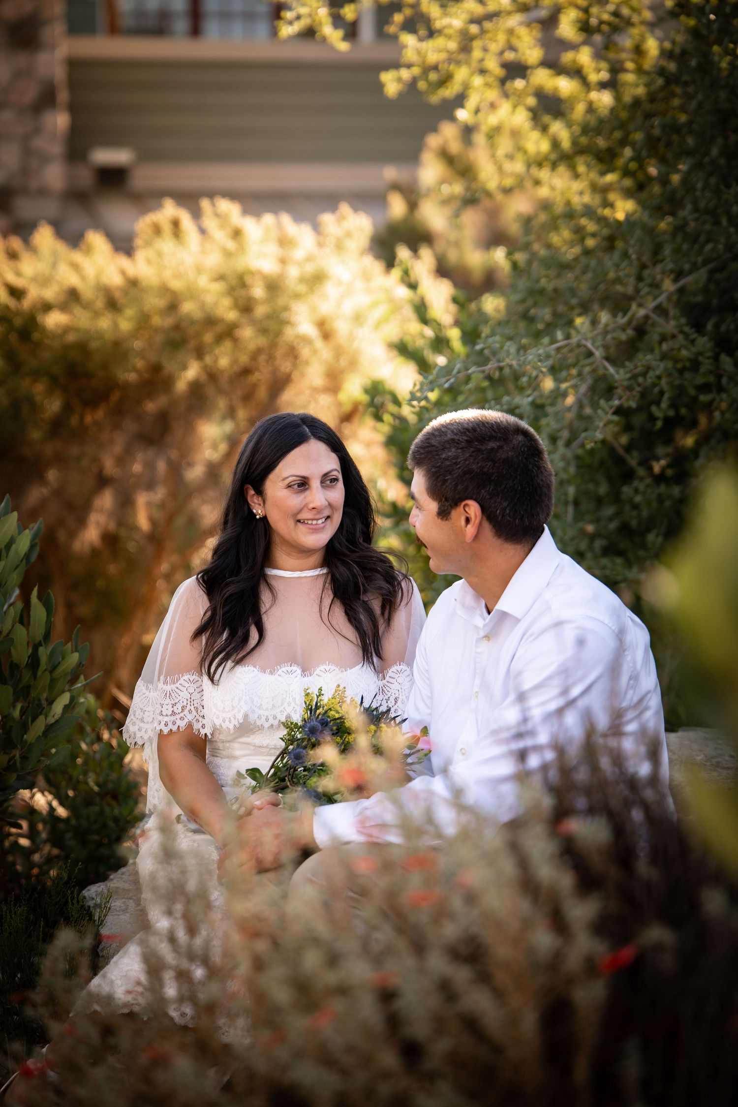 Bride and groom sitting together in a sunlit garden, smiling at each other during wedding portraits.