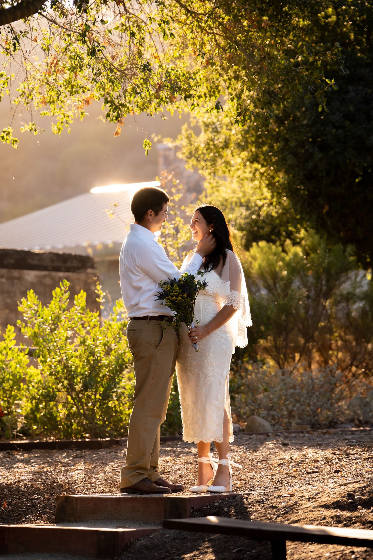 A couple shares a romantic moment at golden hour, bride holding bouquet in white dress, groom in khakis outdoors.