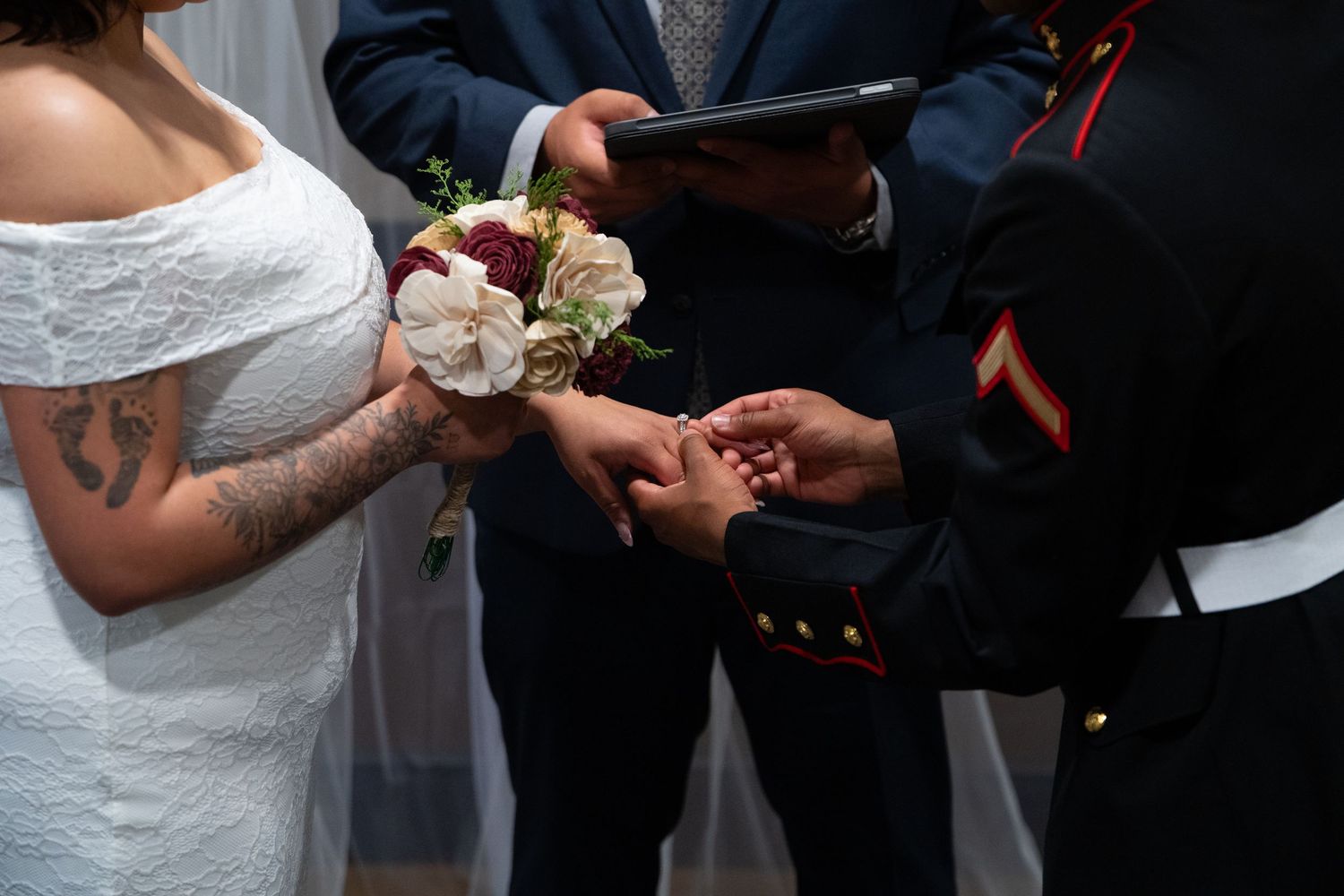 Marine groom placing wedding ring on bride's finger during ceremony, bride holding white and red bouquet.