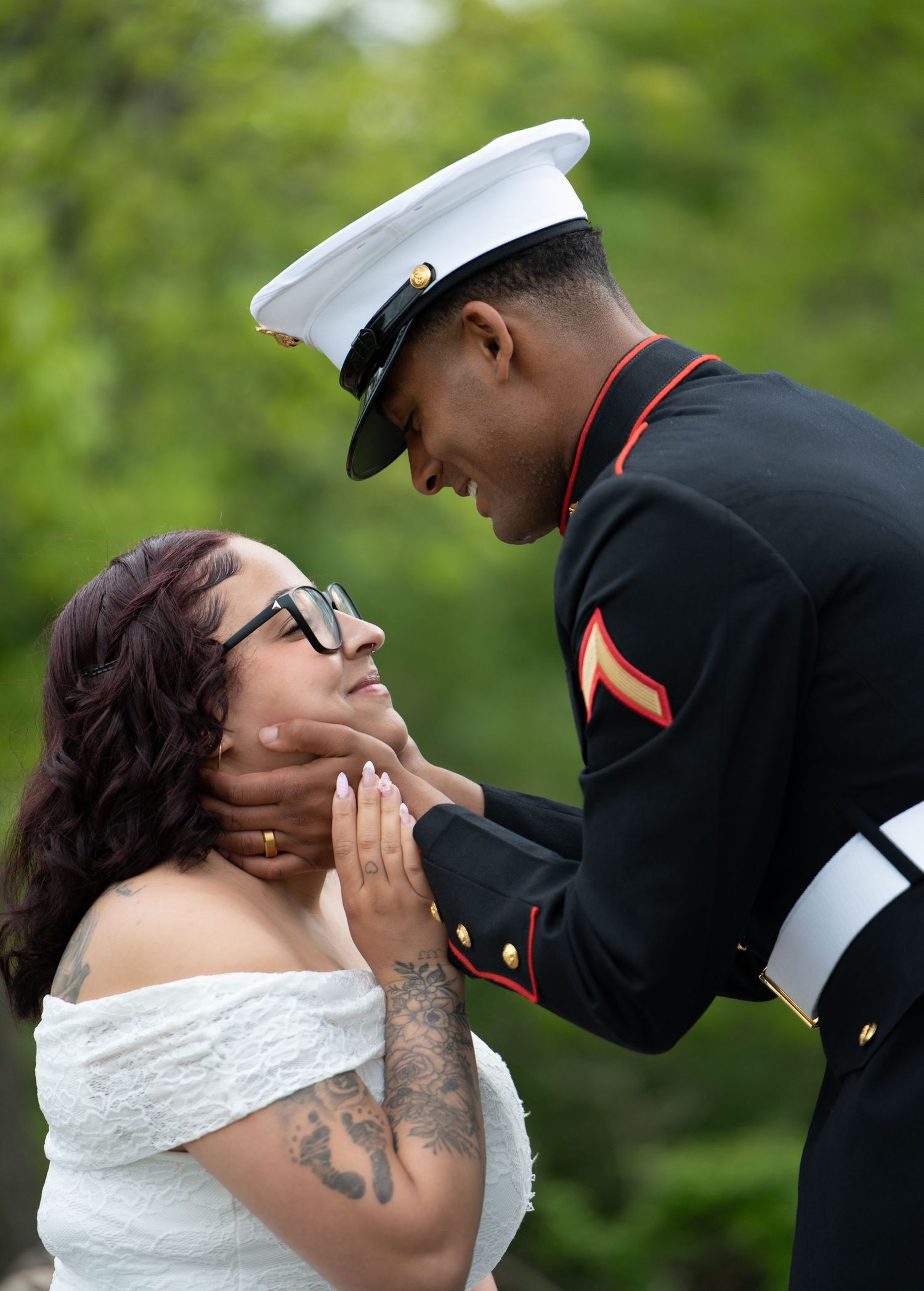 A US Marine in dress blues tenderly holds a woman's face in an outdoor wedding portrait.