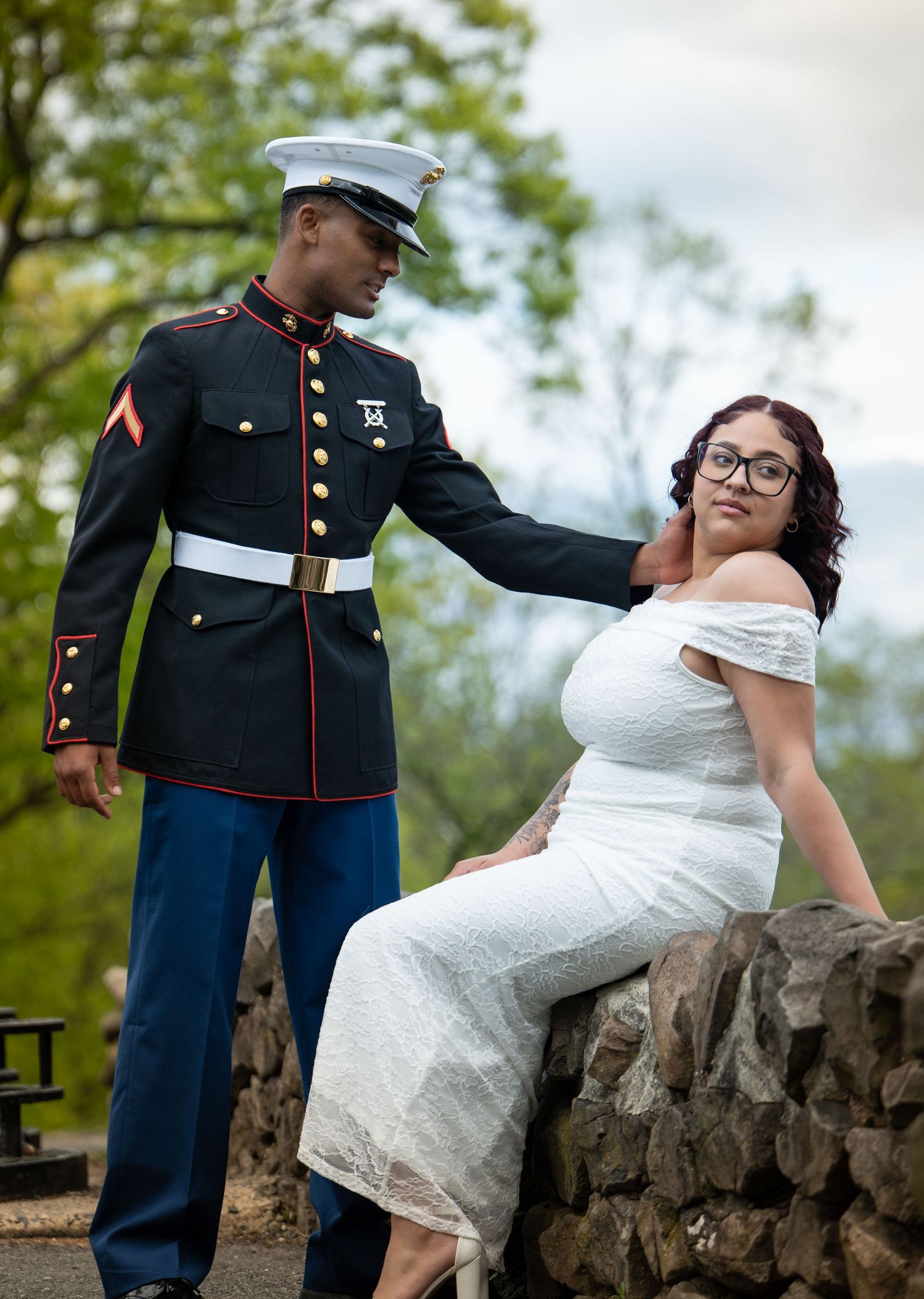 Marine in dress uniform stands beside a woman in white lace dress seated on stone wall outdoors.