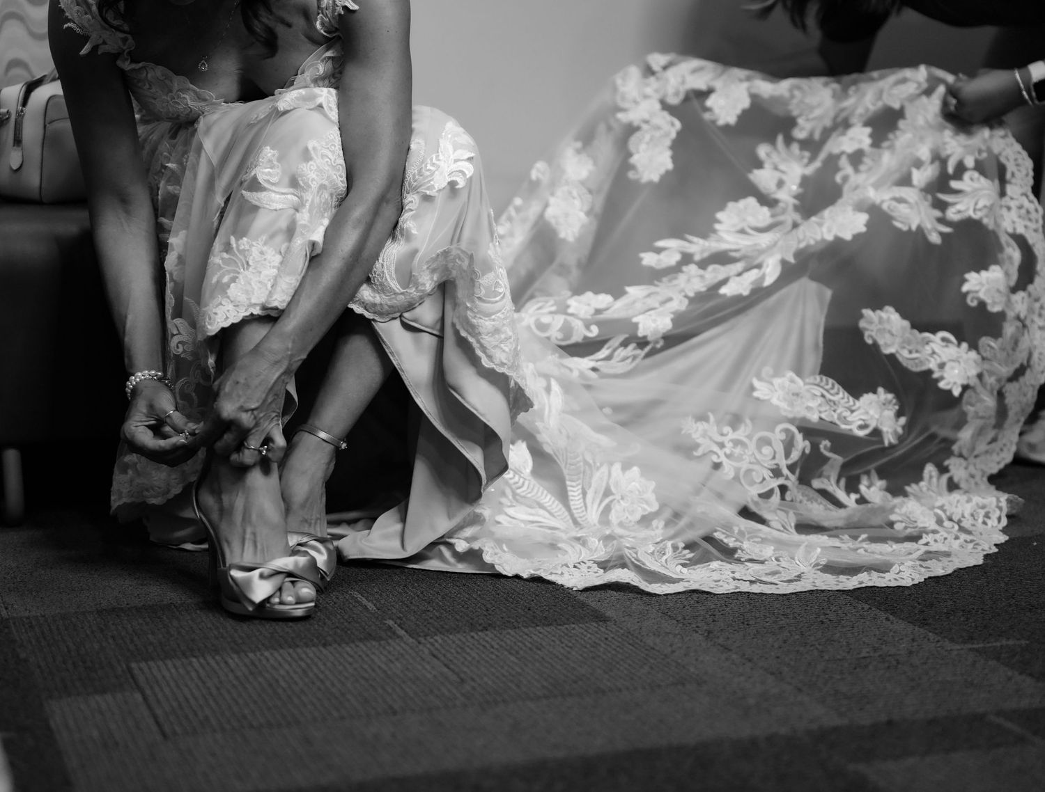 Black and white close-up of a bridesmaid fastening the straps of a brides elegant wedding heels.