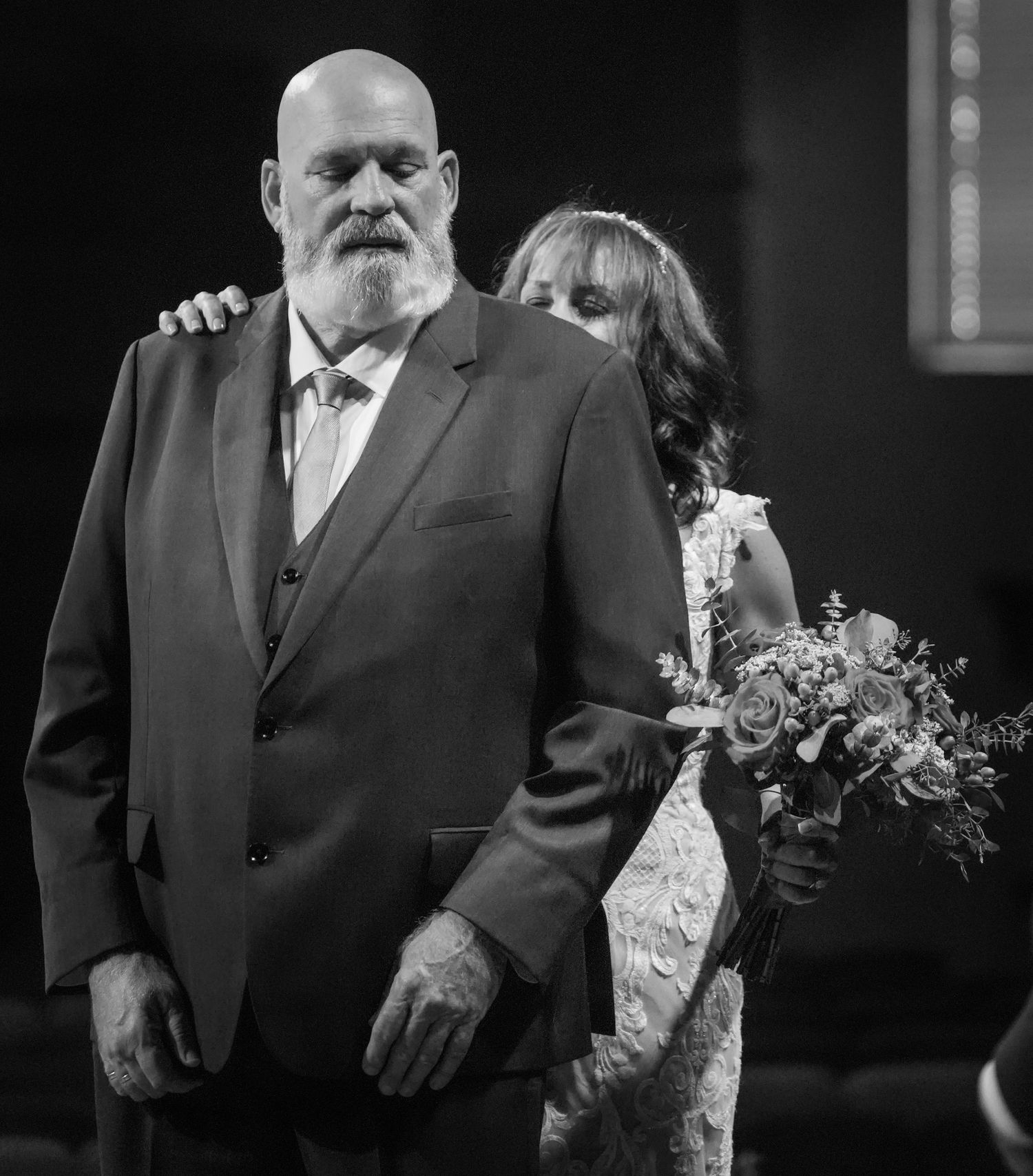 Black and white photo of a bearded man in a suit walking a bride down the aisle at a wedding ceremony.