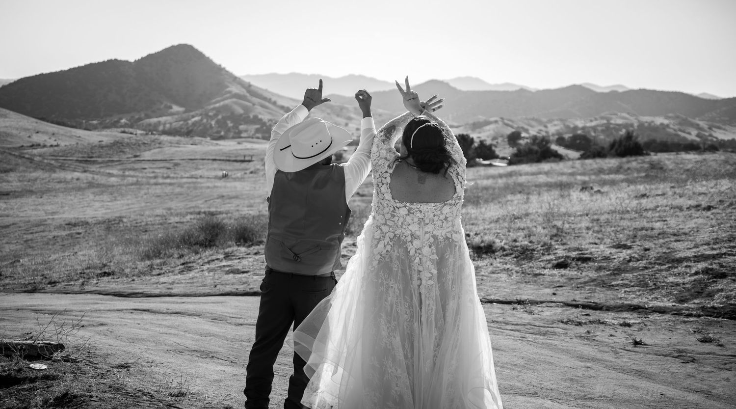 Black and white photo of a bride and groom walking hand in hand through a desert landscape with mountains in the background.
