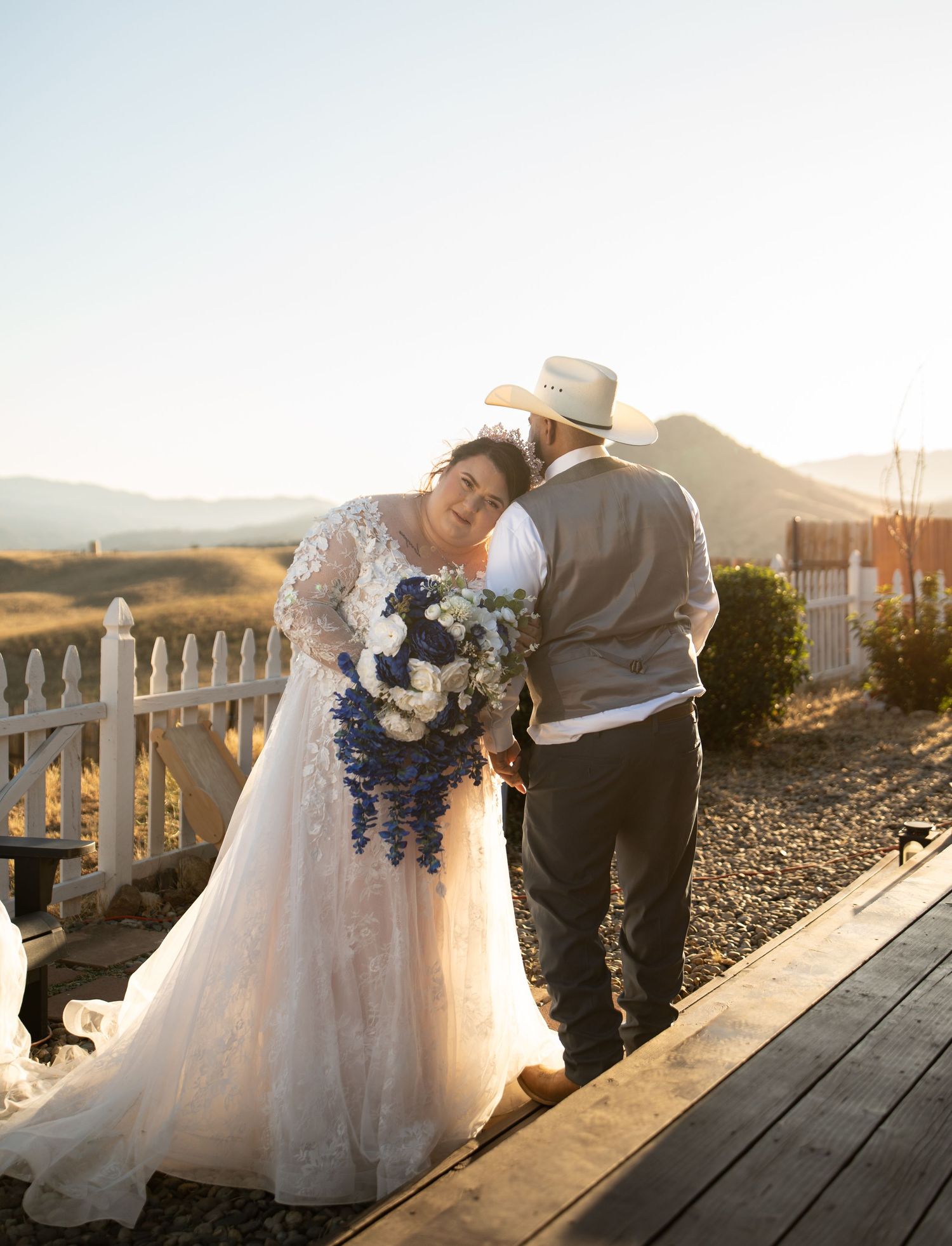 Bride in lace gown holding blue bouquet shares romantic moment with groom in cowboy hat on rustic deck at sunset.