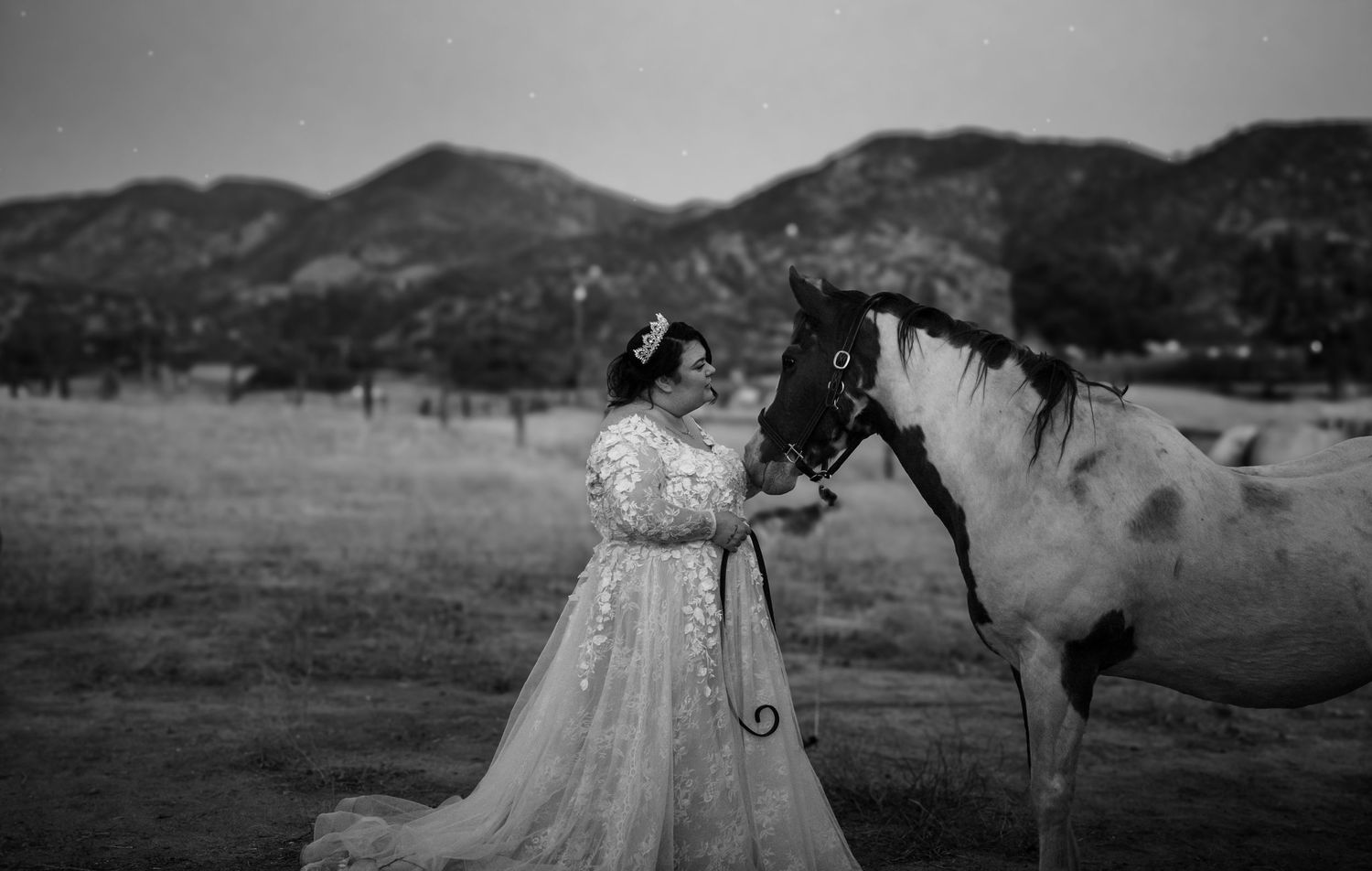 Black and white photo of a young woman in a white gown posing with a paint horse in a mountain field.
