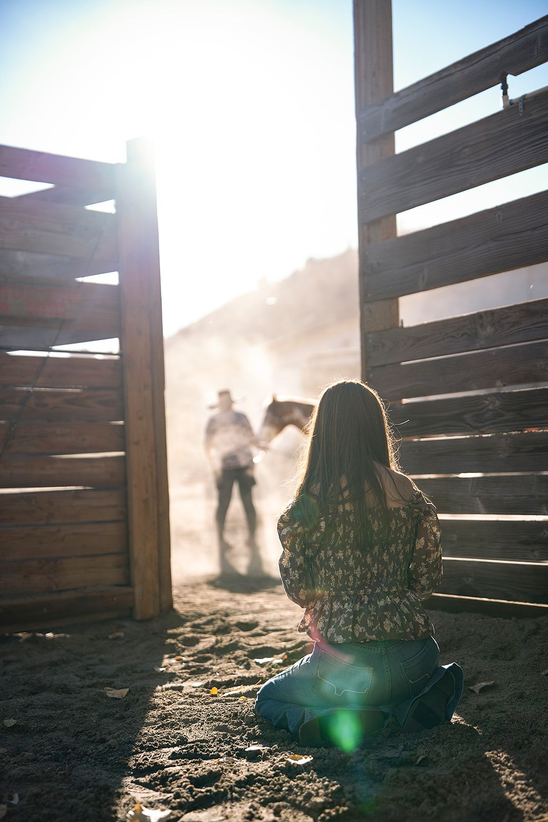 Girl sitting in barn doorway watching cowboy lead horse through dusty sunlit corral.