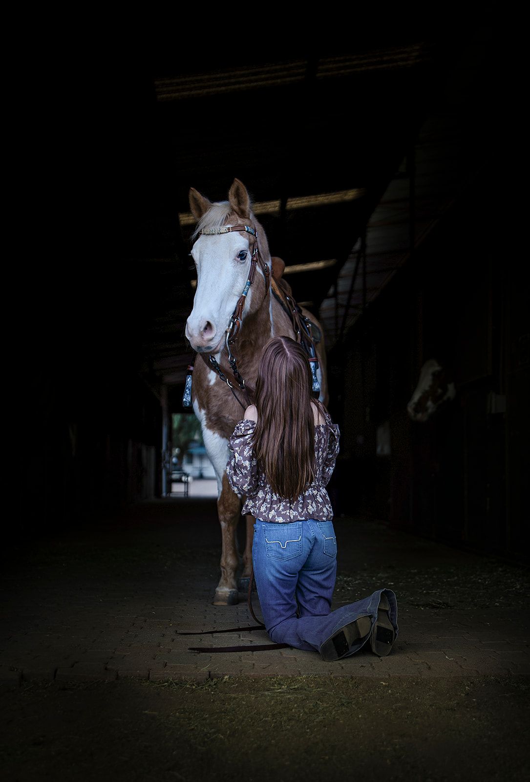 Young woman kneeling beside white and brown horse in dark stable interior.