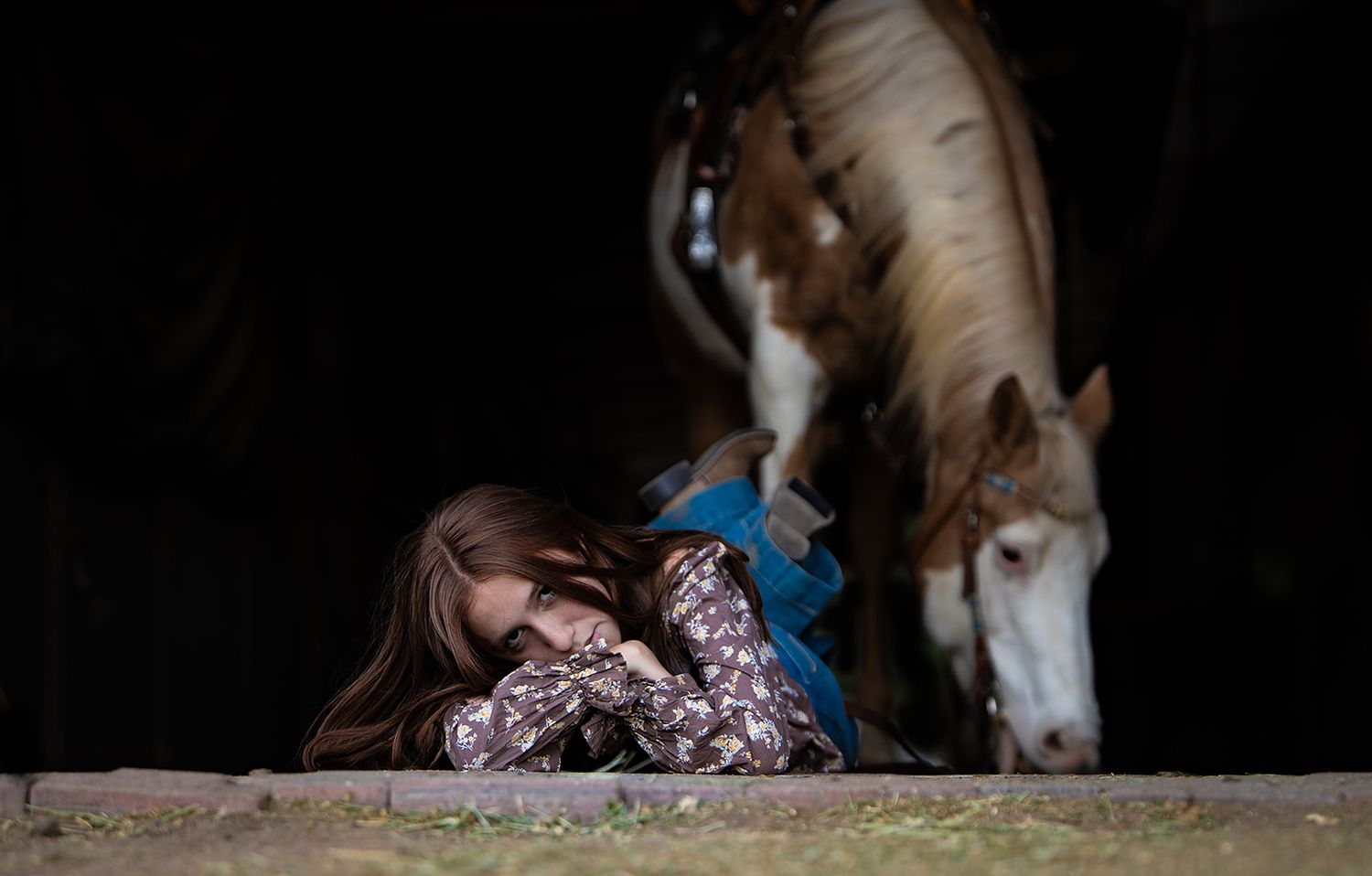 Girl lying on barn floor beside a paint horse grazing in rustic stable.