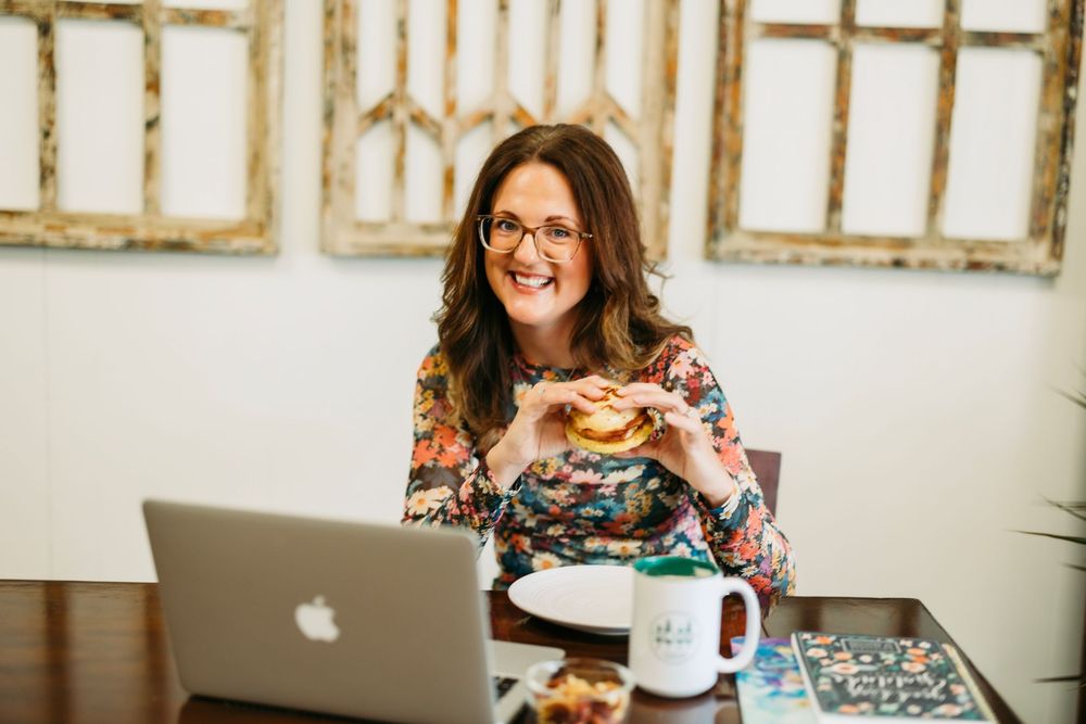 Woman in floral dress smiles while working on laptop at desk with coffee and sandwich.