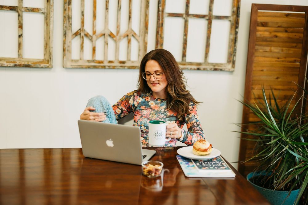 A woman in glasses sits at a wooden table working on a laptop in a bright interior space.