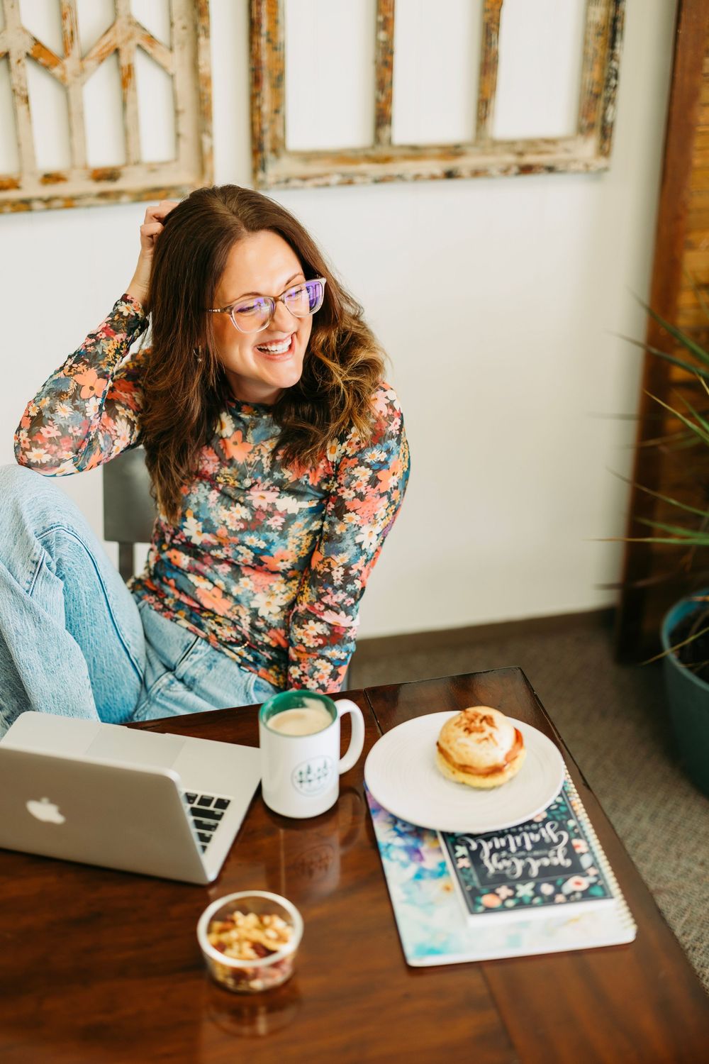 A person in a floral top works on a laptop at a desk with coffee and pastry.
