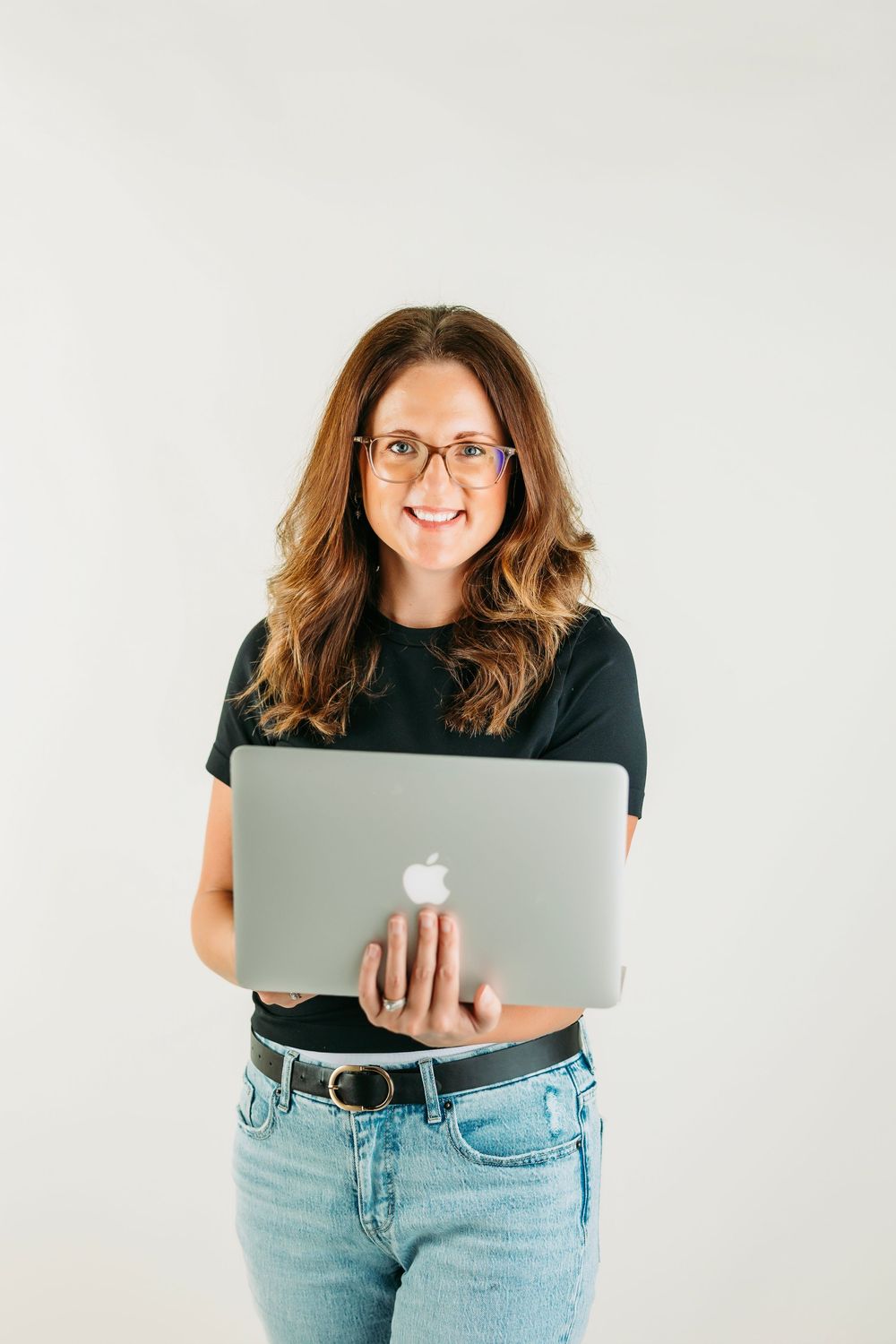 Person in glasses and casual attire holding a MacBook laptop while standing against white background.