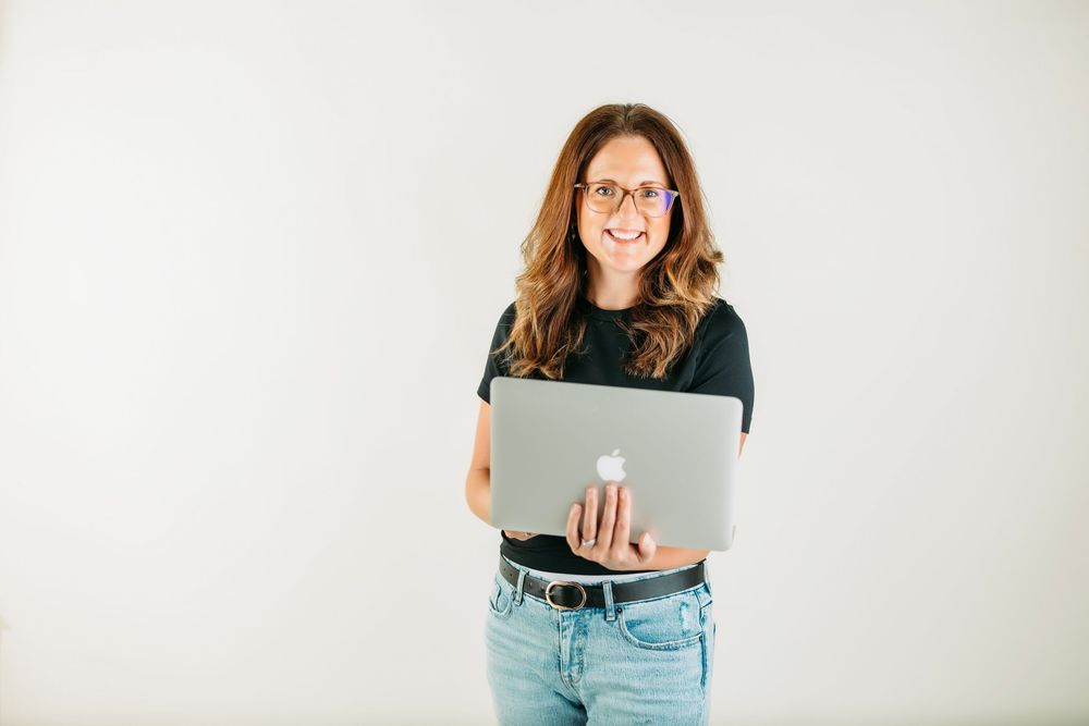 Professional wearing glasses and casual attire holds laptop while standing against white background.