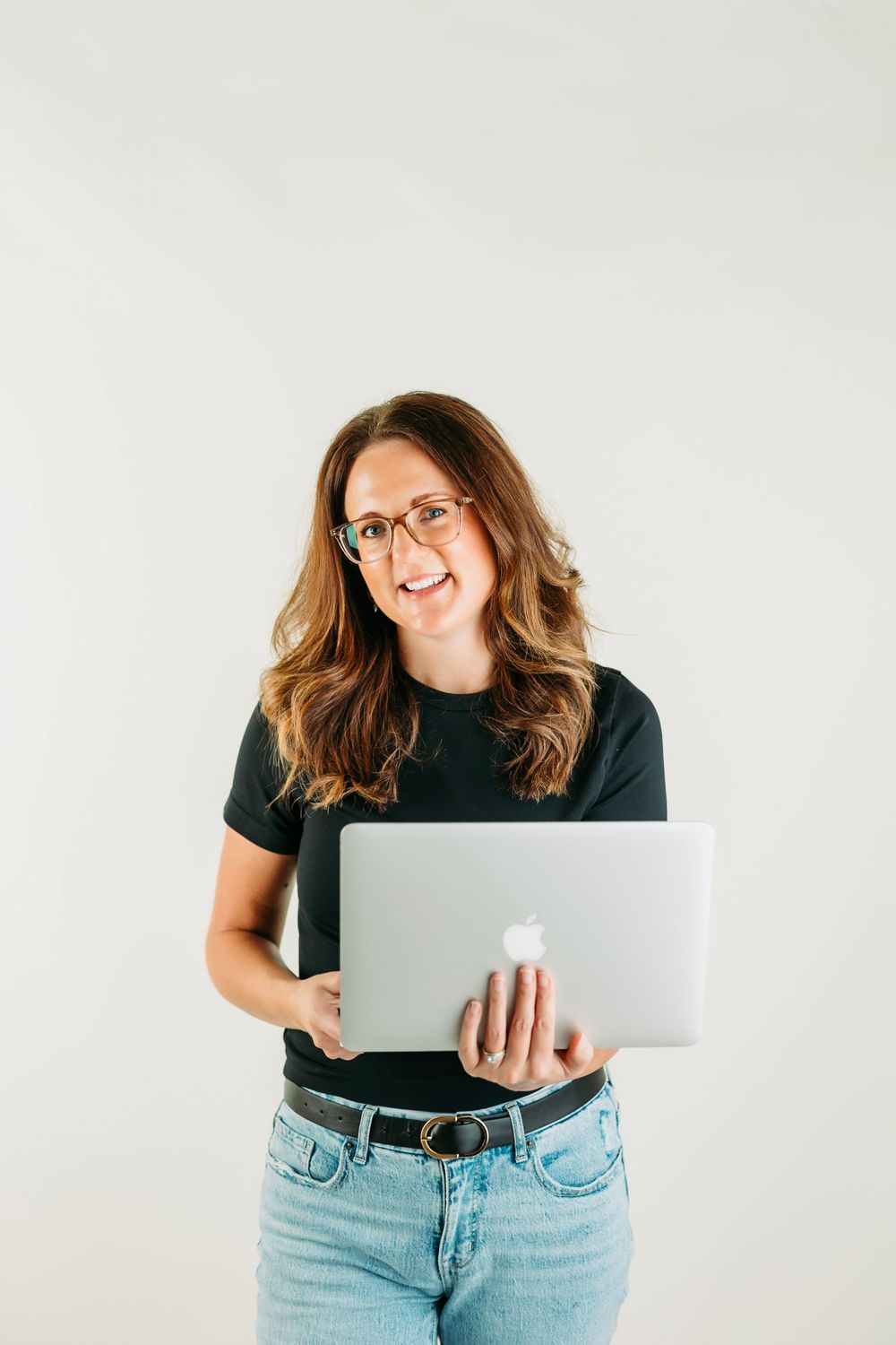 Person in black shirt and jeans poses casually with laptop against white backdrop.