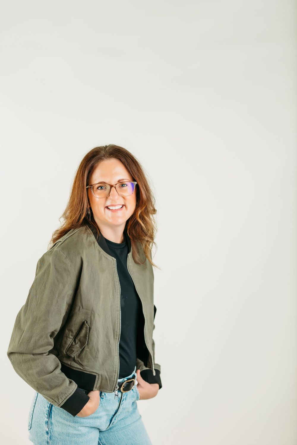 Professional wearing olive jacket and glasses poses against white background in casual business attire.