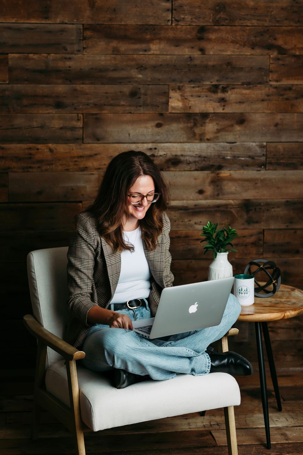 A person in a blazer and jeans works on a MacBook while sitting in a white armchair against a rustic wooden wall.