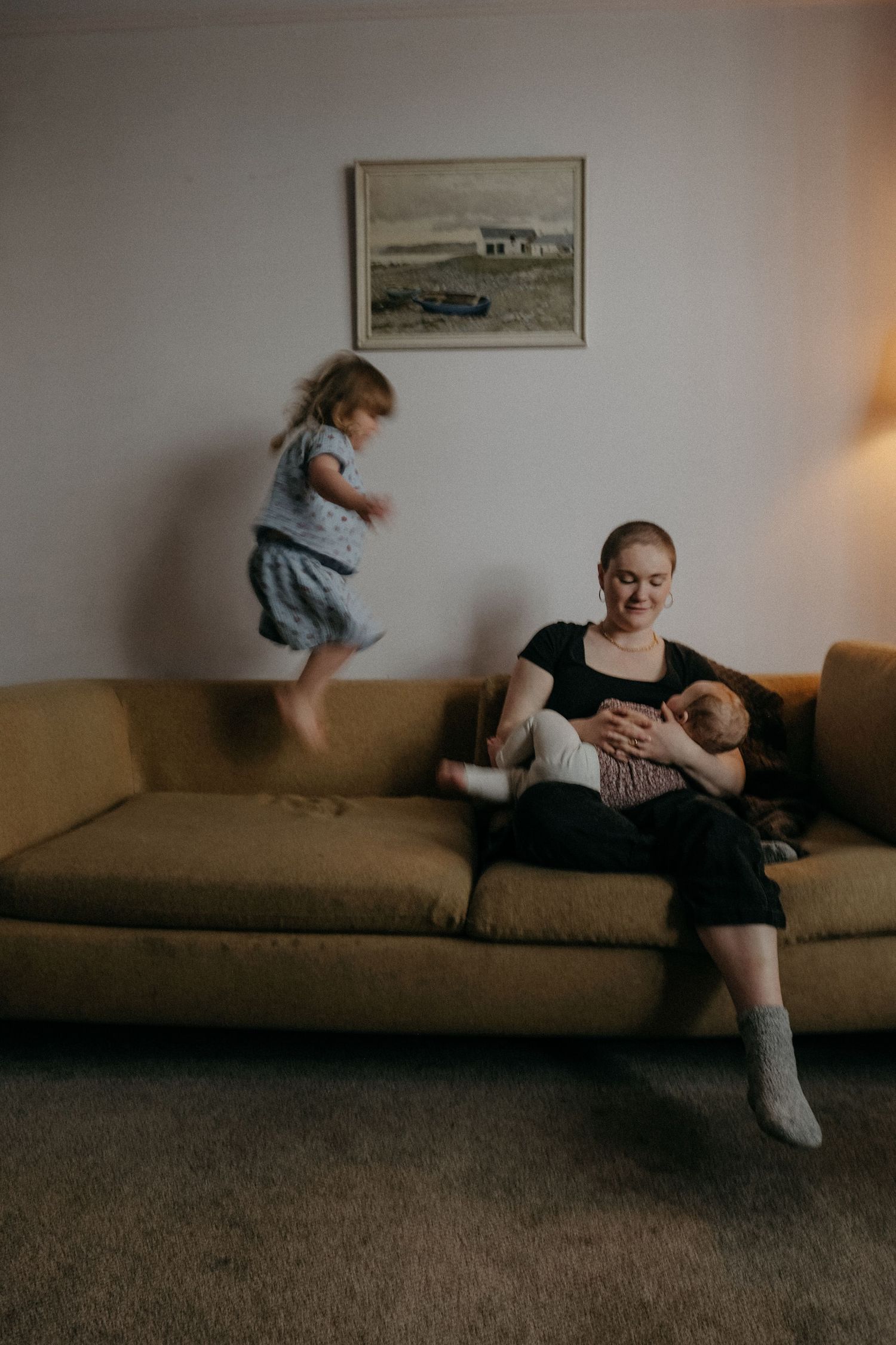 Mother sitting on sofa holding baby while toddler jumps beside him during a relaxed in-home lifestyle family photography session in heaton