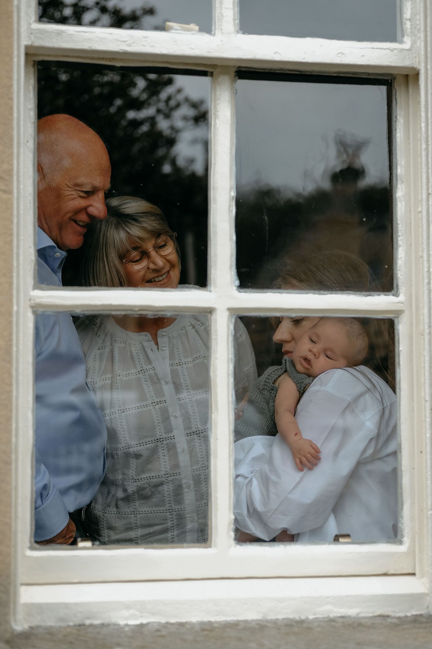 Grandparents and daughter sharing a moment together while standing by a window during an in-home newborn photography session in Corbridge